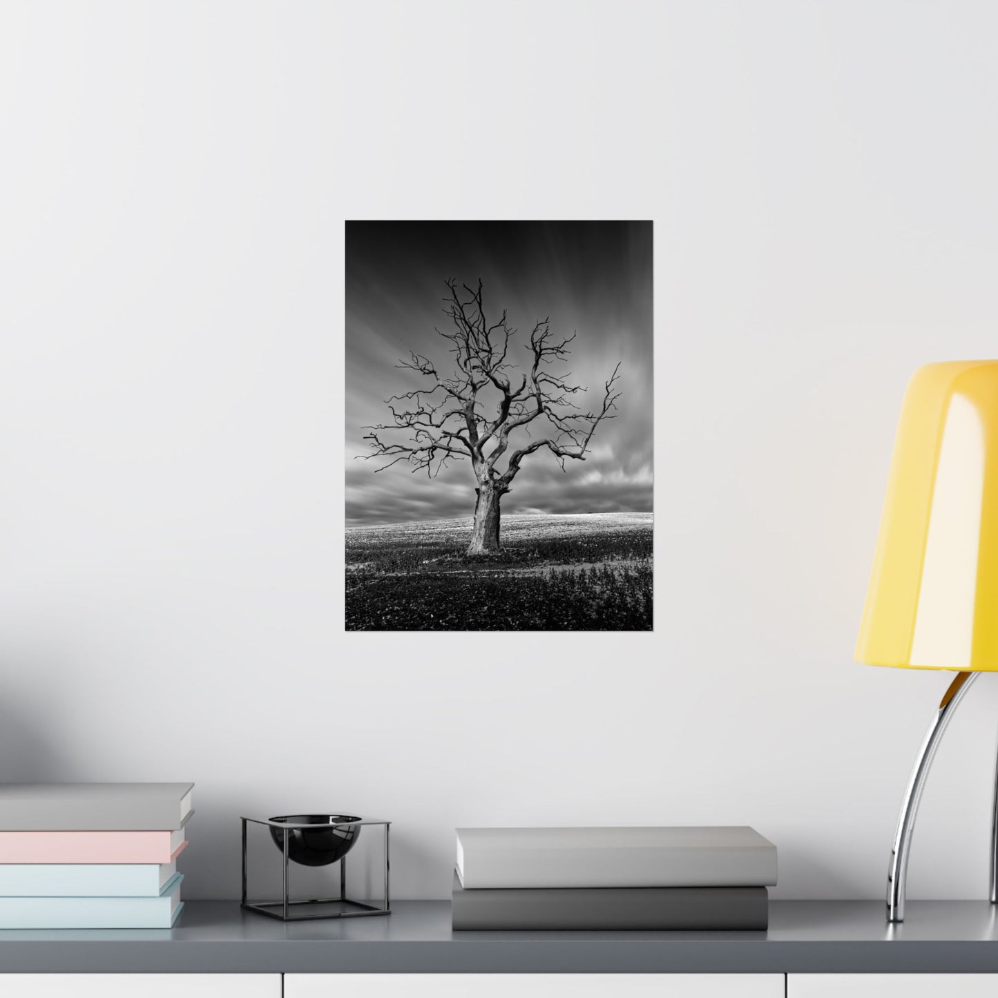 Framed black and white print of a tree on a wall above a desk with books and a lamp.