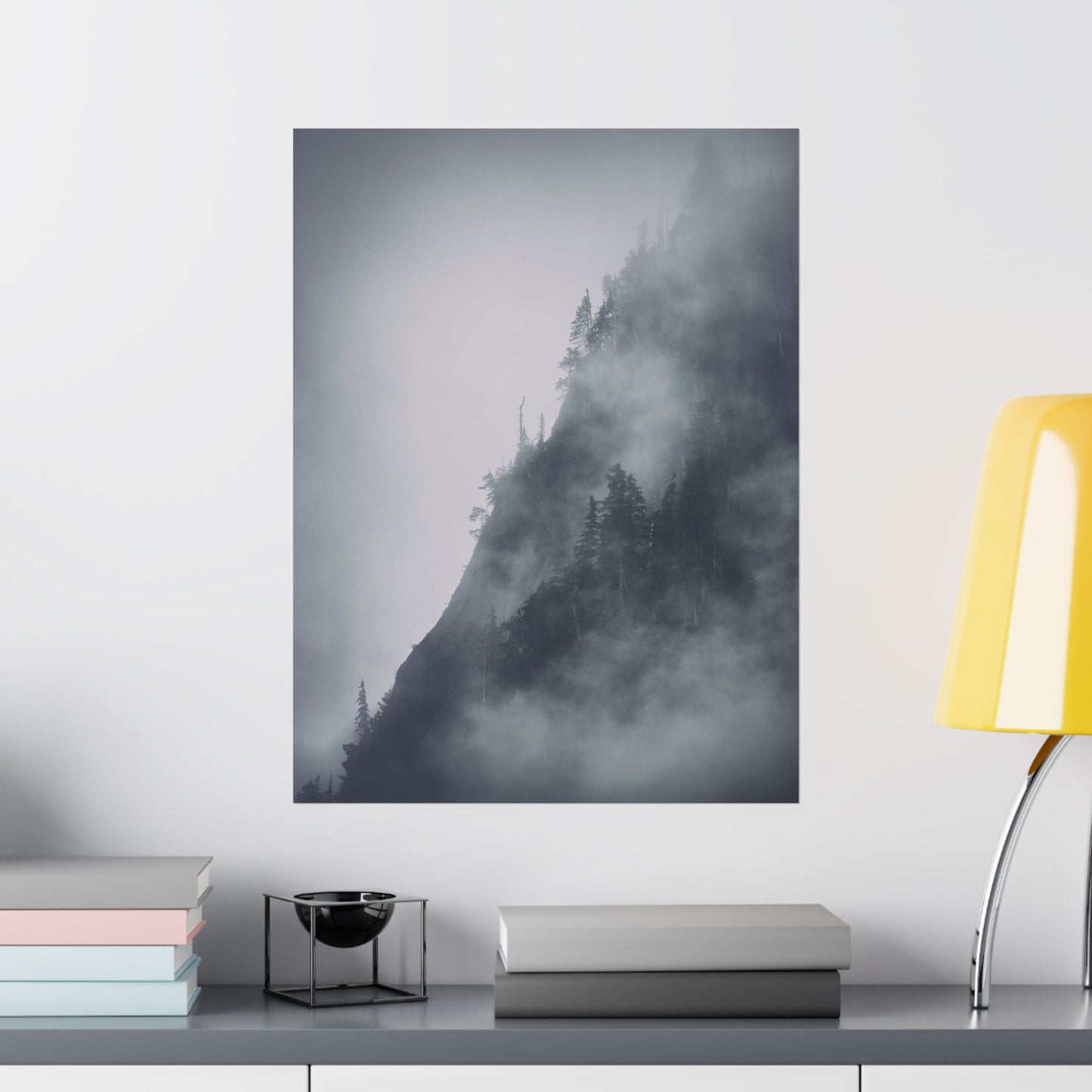 Foggy mountain landscape print on a wall above a shelf with books and a lamp.