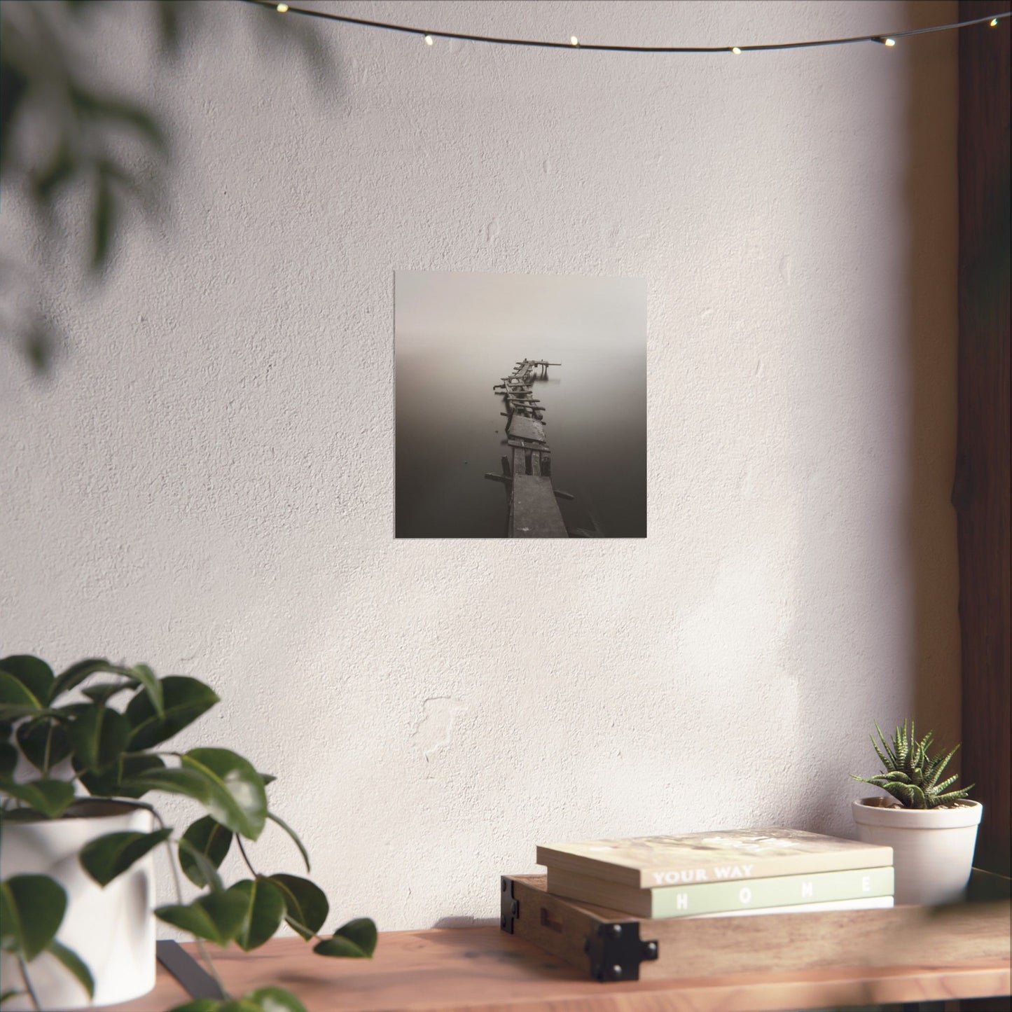 Framed artwork of a lighthouse on a white wall above a wooden desk with plants and books.