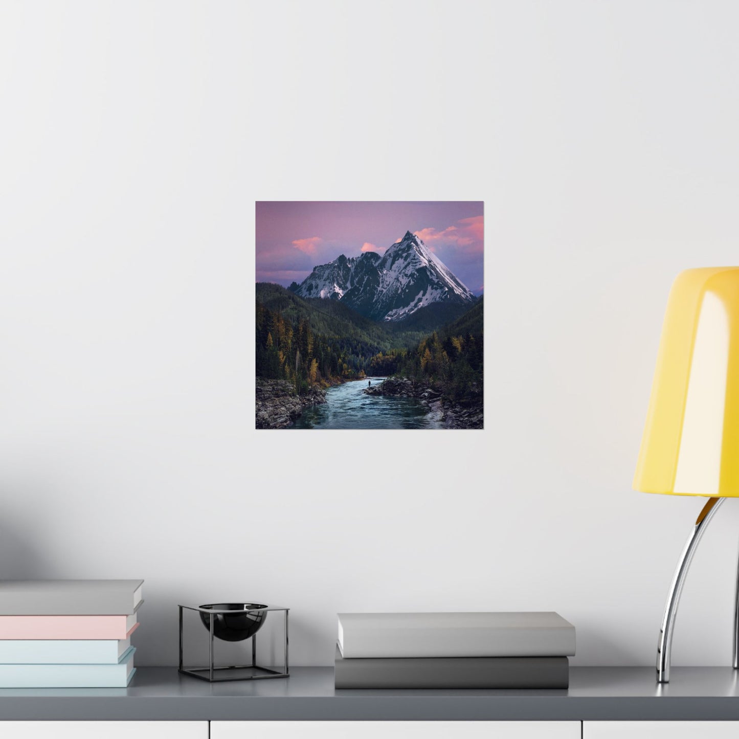 Framed mountain landscape print on a wall above a desk with books and a lamp.