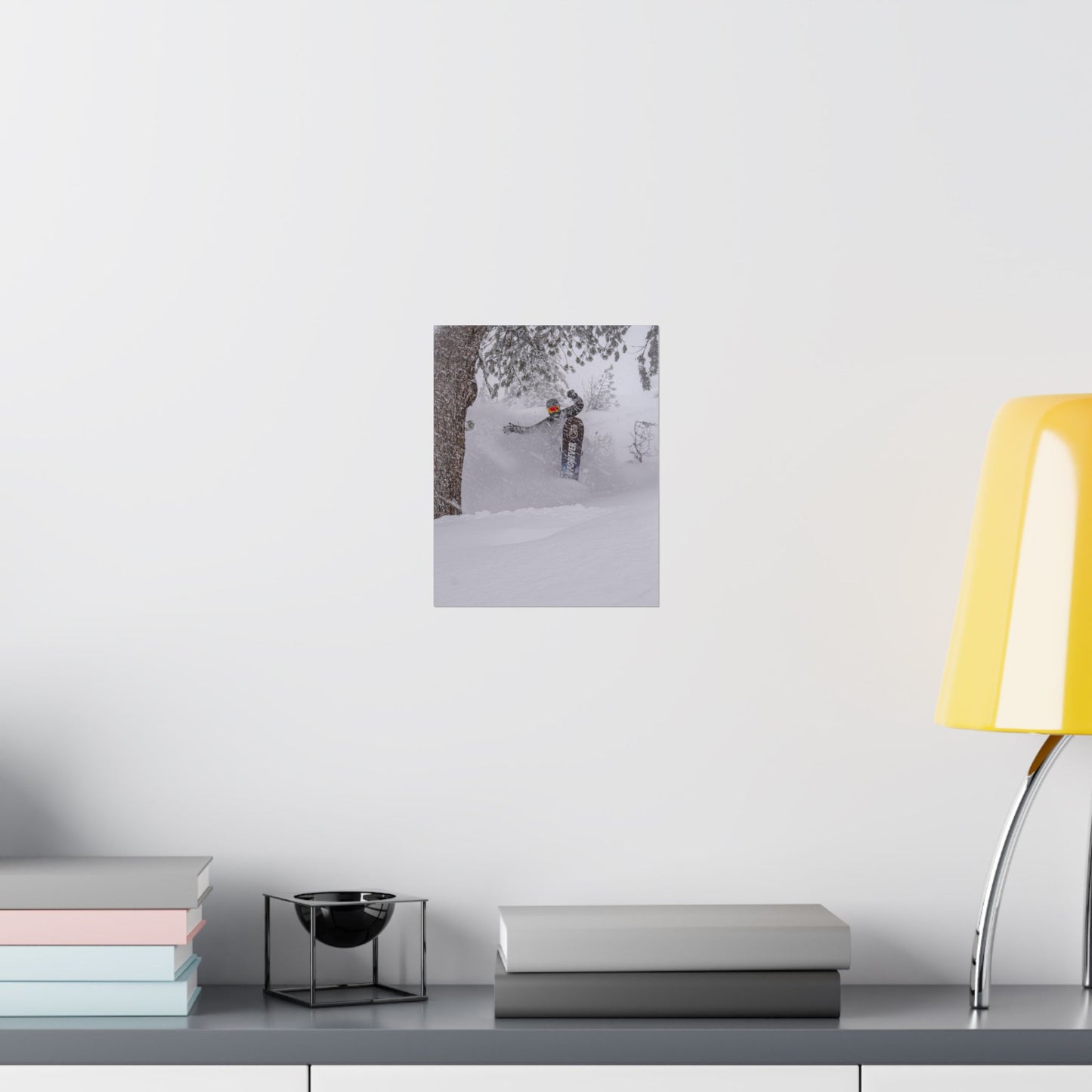 Framed photograph of a person skiing on a snowy landscape hanging on a white wall above a desk with books and a lamp.