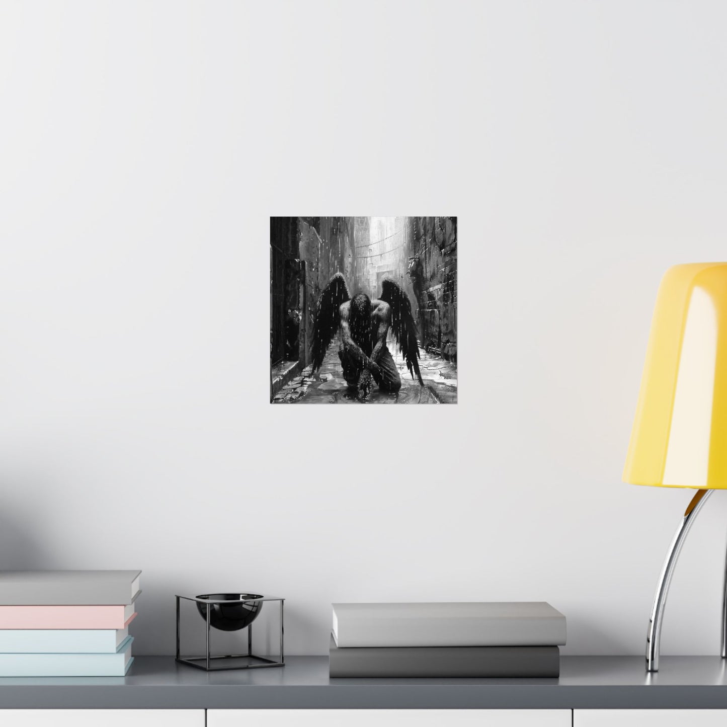Framed black and white artwork of two figures in a forest on a wall above a shelf with books and a lamp.