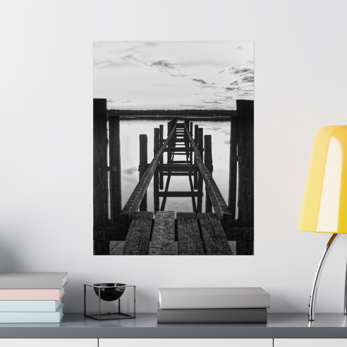 Black and white photo of a dock on a wall above a shelf with books and a lamp.