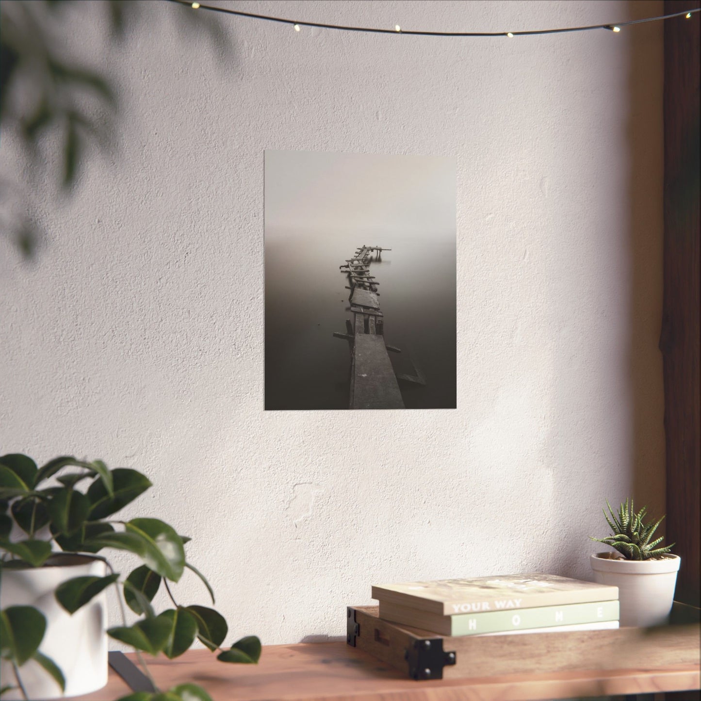 Poster of a misty dock scene on a wall above a wooden table with plants and books.