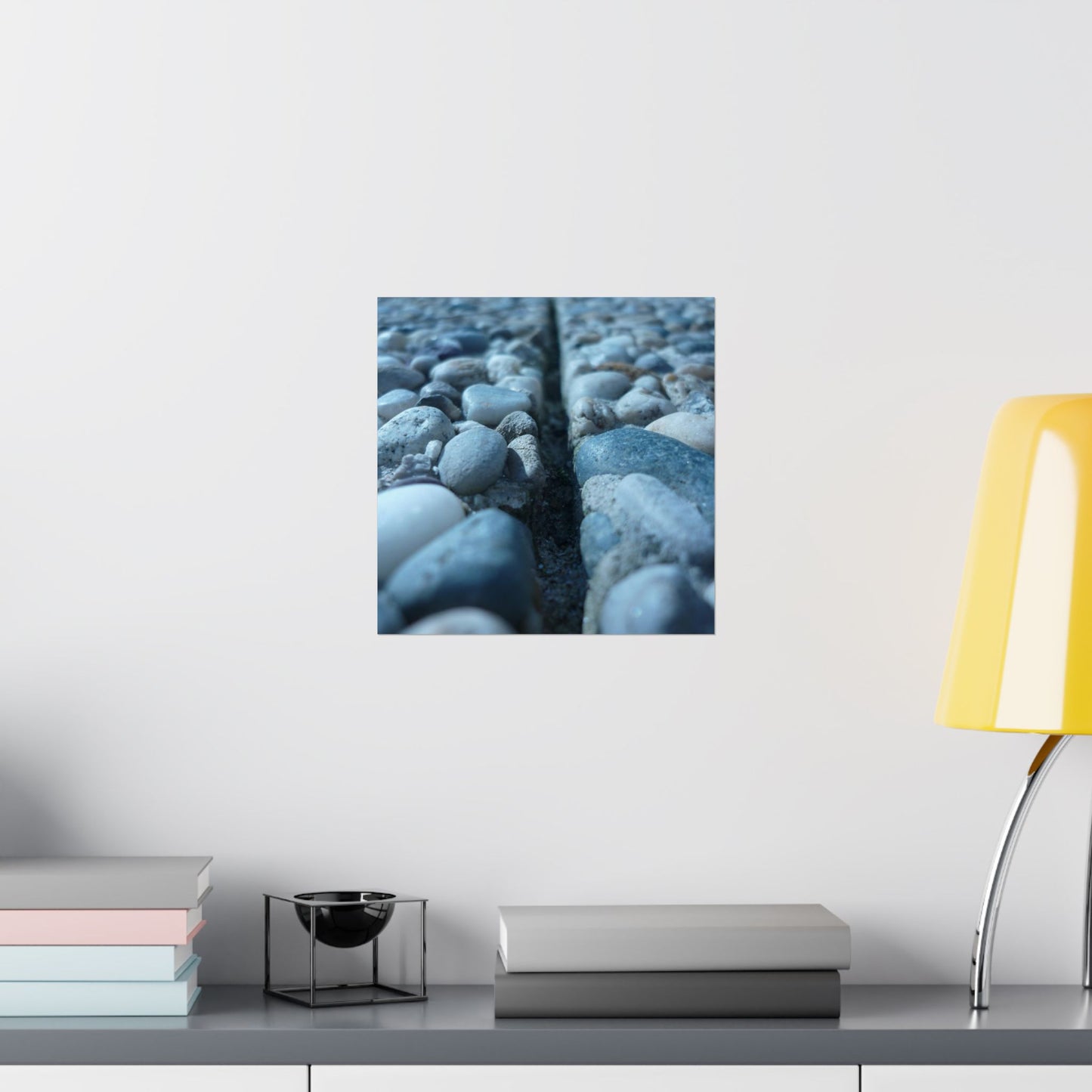 Wall art of pebbles on a white wall above a shelf with books and a lamp.