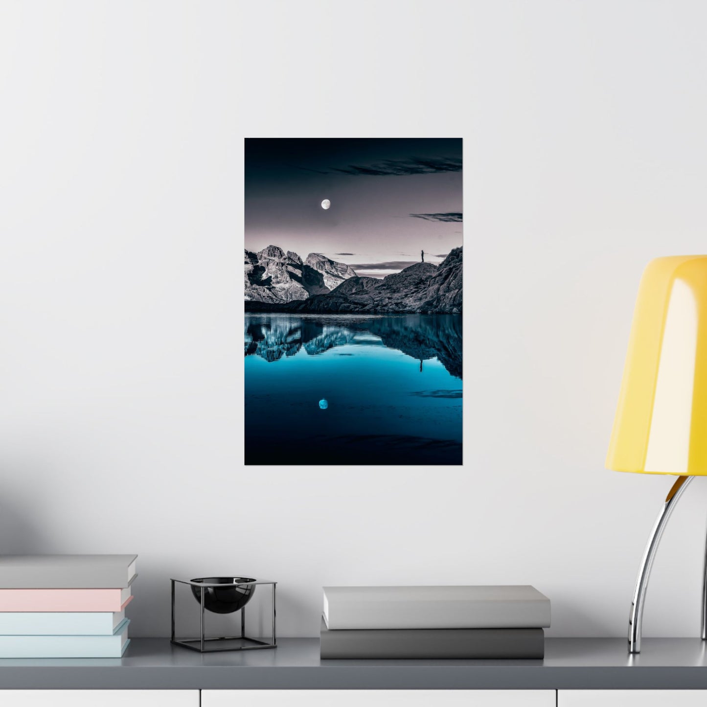 Framed mountain landscape print on a wall above a shelf with books and a lamp.