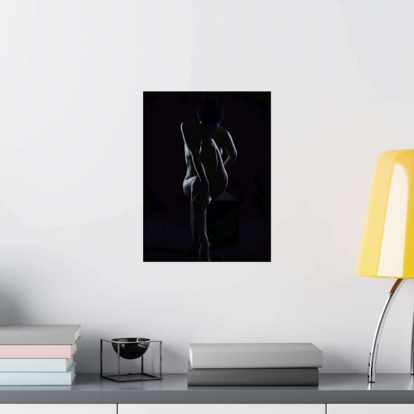 Framed black and white art print of a nude figure on a wall above a shelf with books and a lamp.