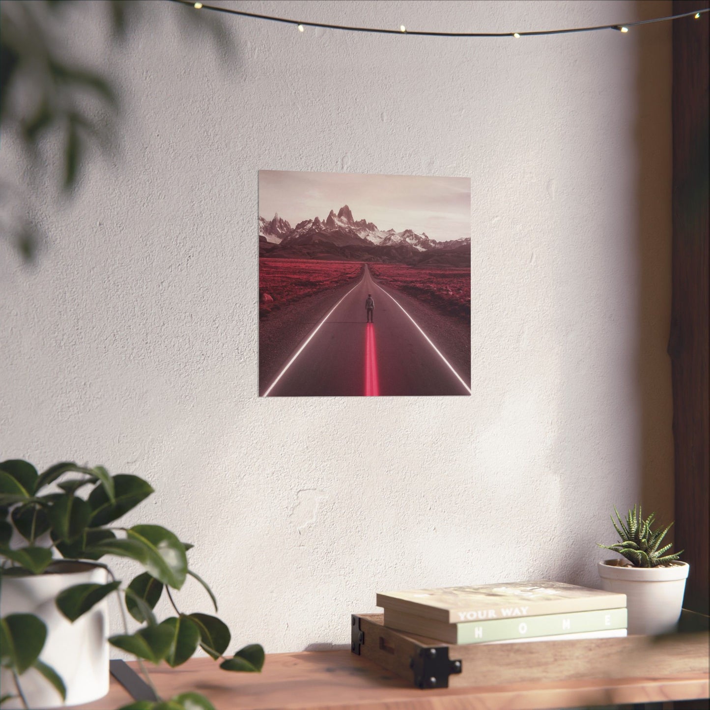 Framed artwork of a road with mountains on a white wall above a wooden desk with plants.