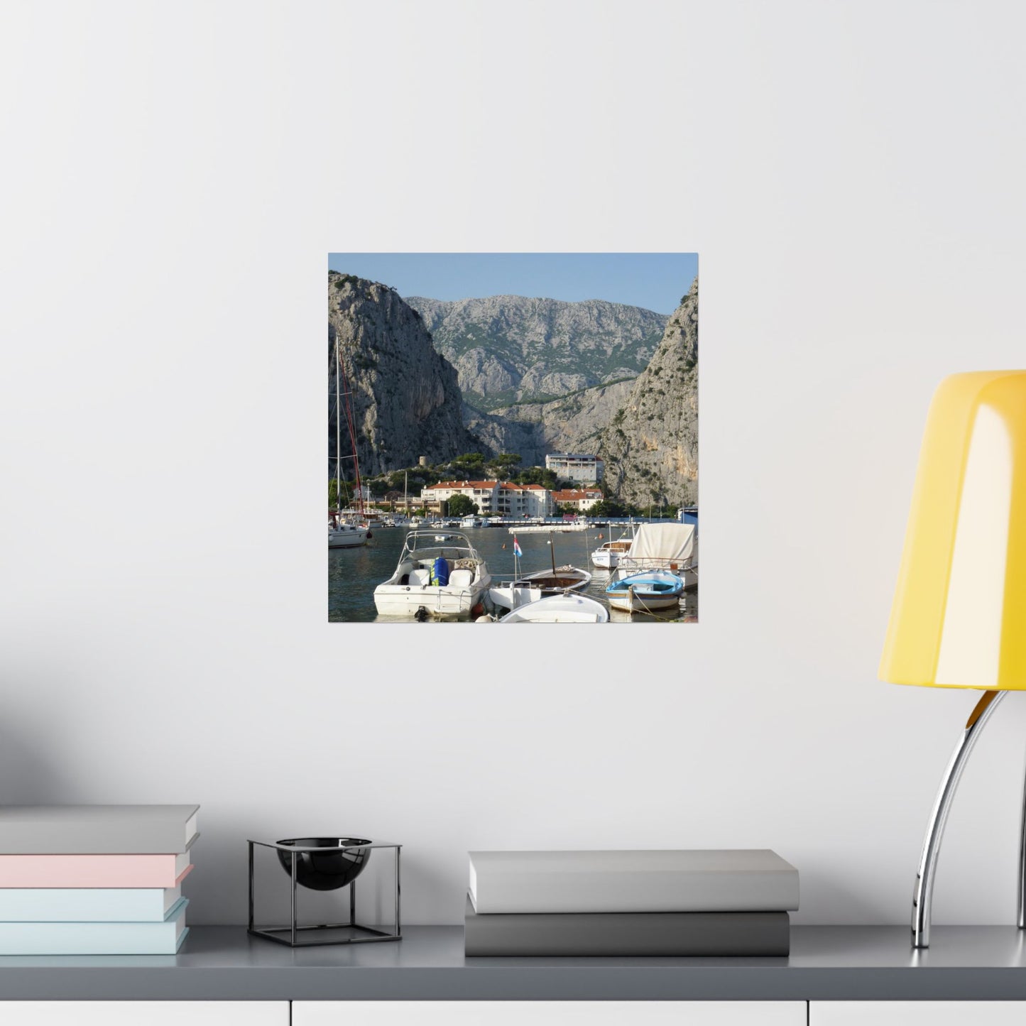 Framed photograph of a coastal scene with boats and mountains on a wall above a shelf.