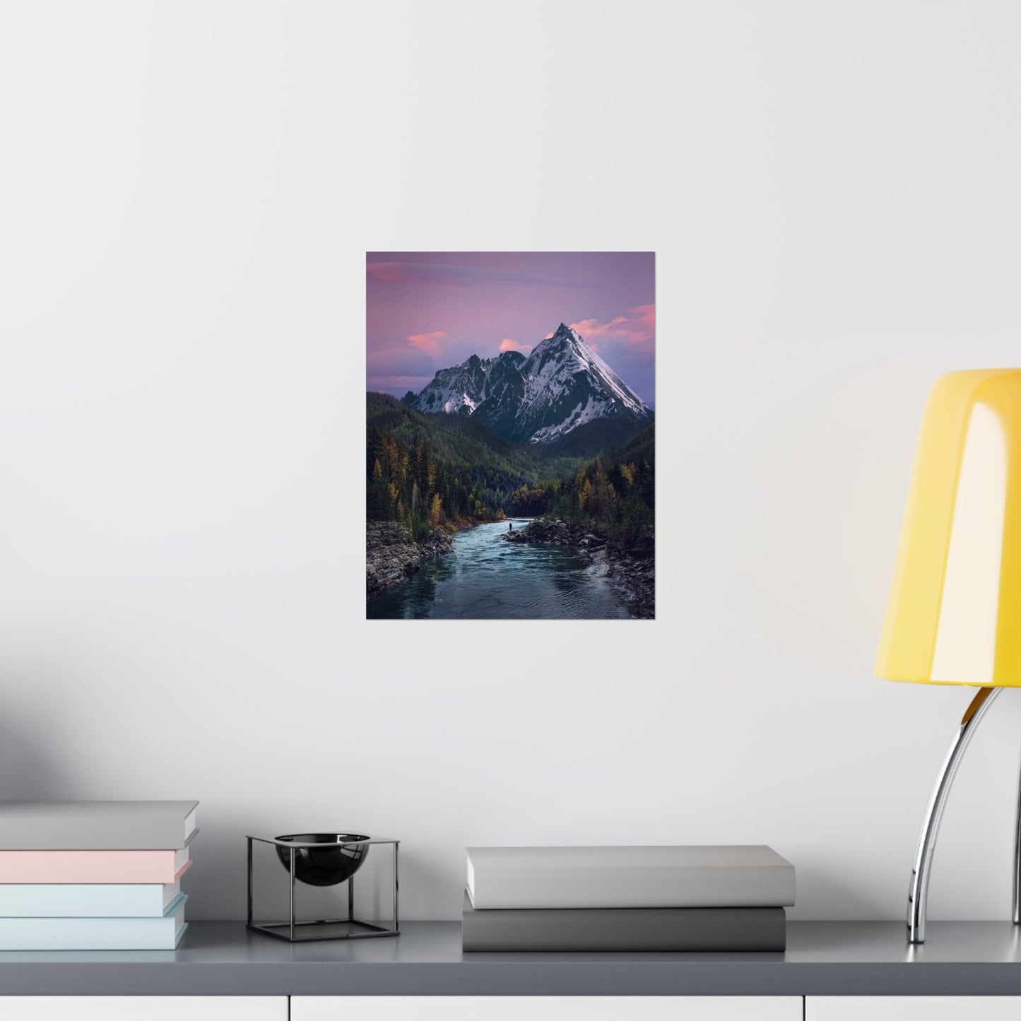 Framed mountain landscape print on a wall above a desk with books and a lamp.