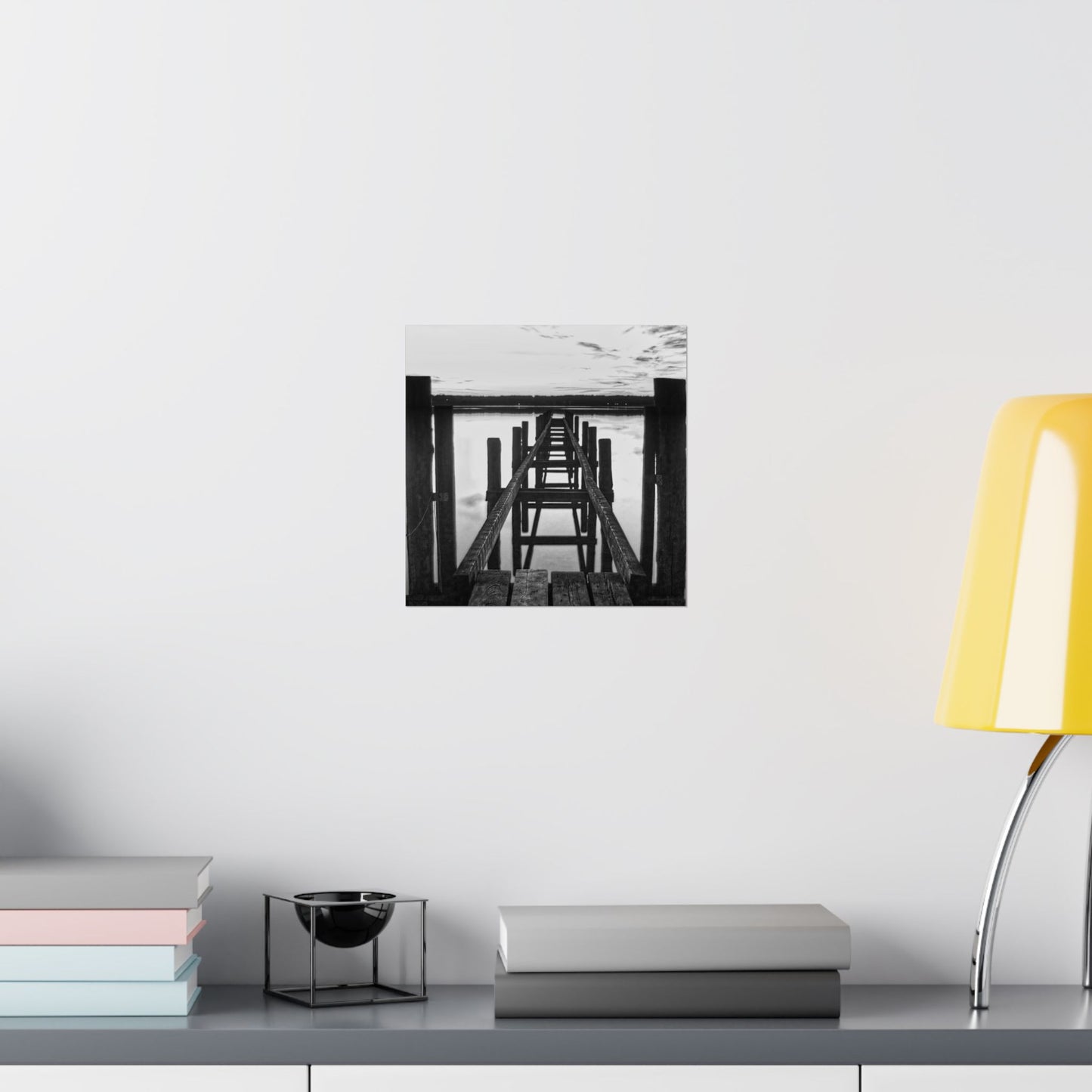 Wall art of a black and white pier on a white wall above a shelf with books and a lamp.