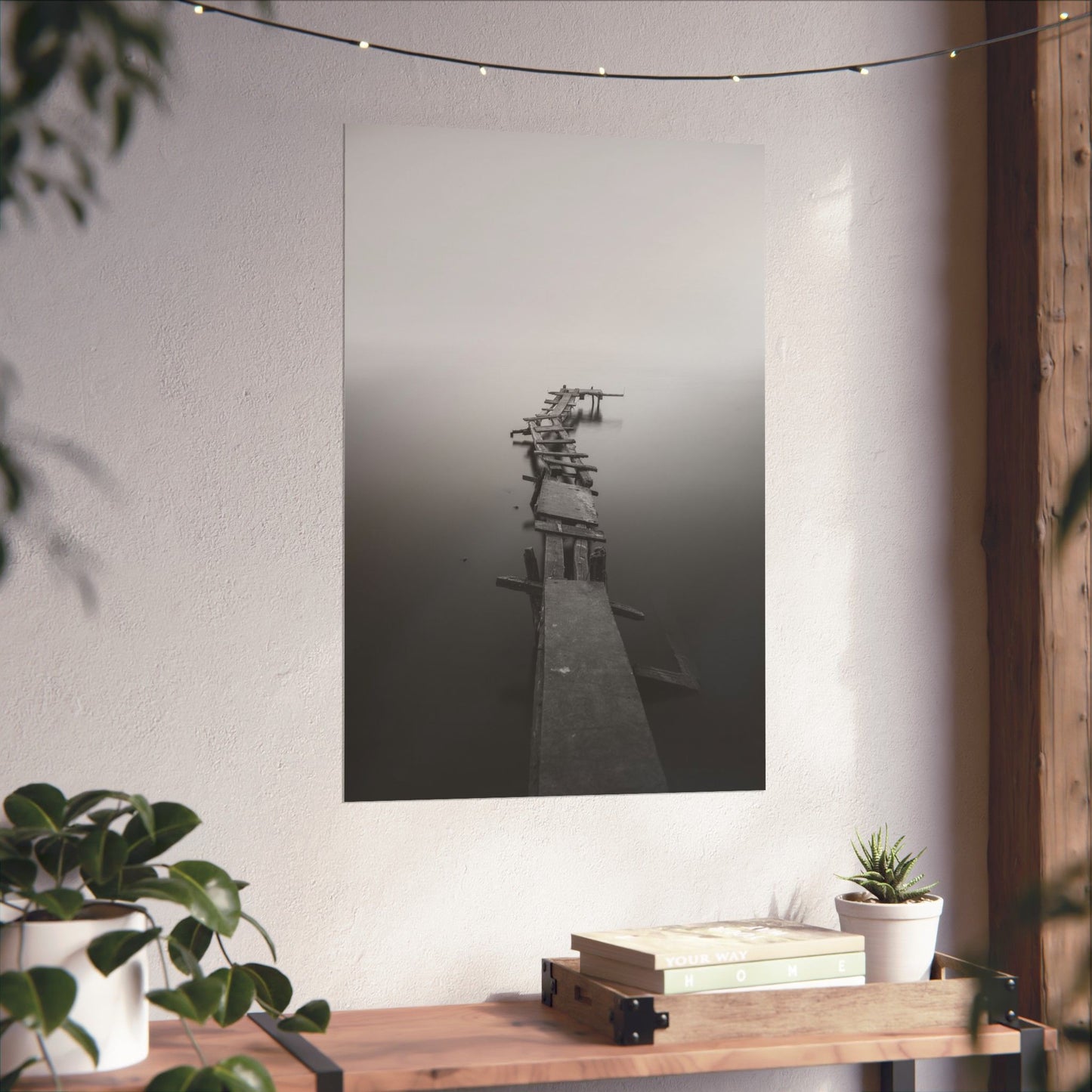 Framed artwork of a dock on a white wall with plants and books below.