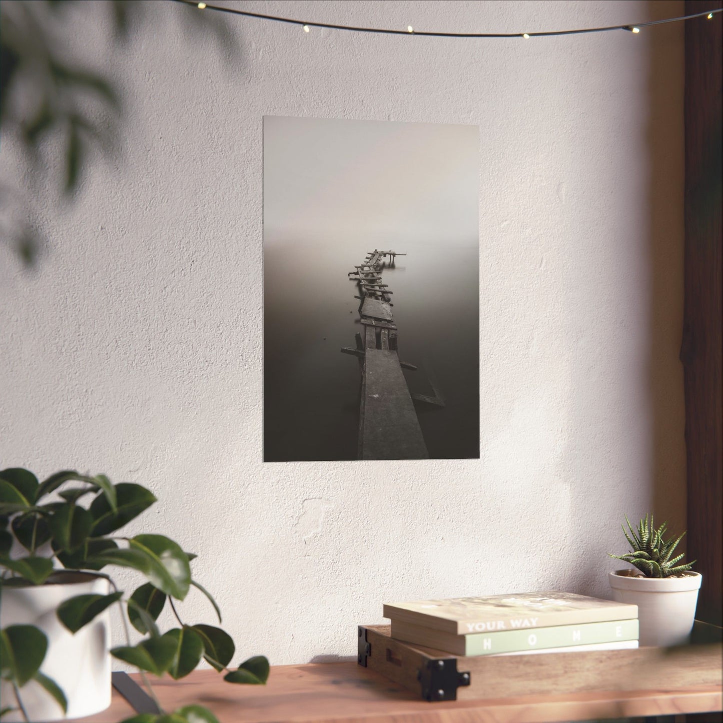 Framed black and white photograph of a dock on a wall above a wooden desk with plants and books.
