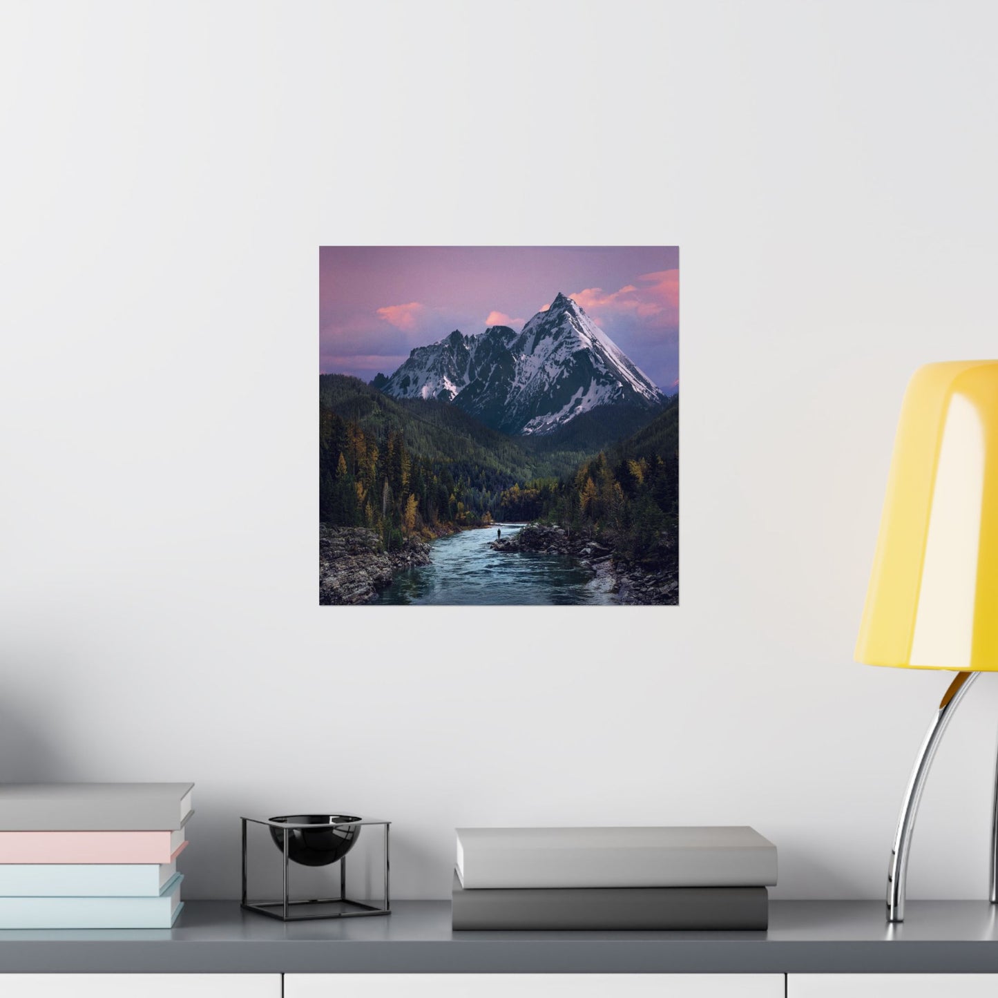 Framed mountain landscape print on a wall above a desk with books and a lamp.