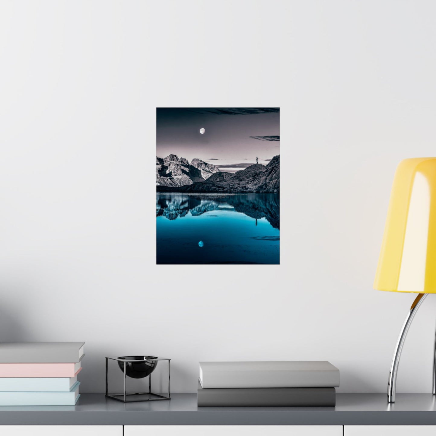 Framed landscape print of a mountain and lake scene on a wall above a desk with books and a lamp.