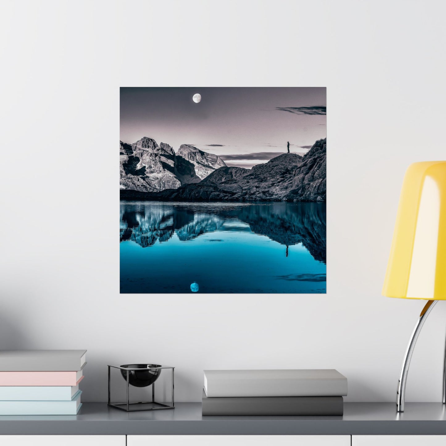 Framed mountain landscape print on a wall above a shelf with books and decor items.