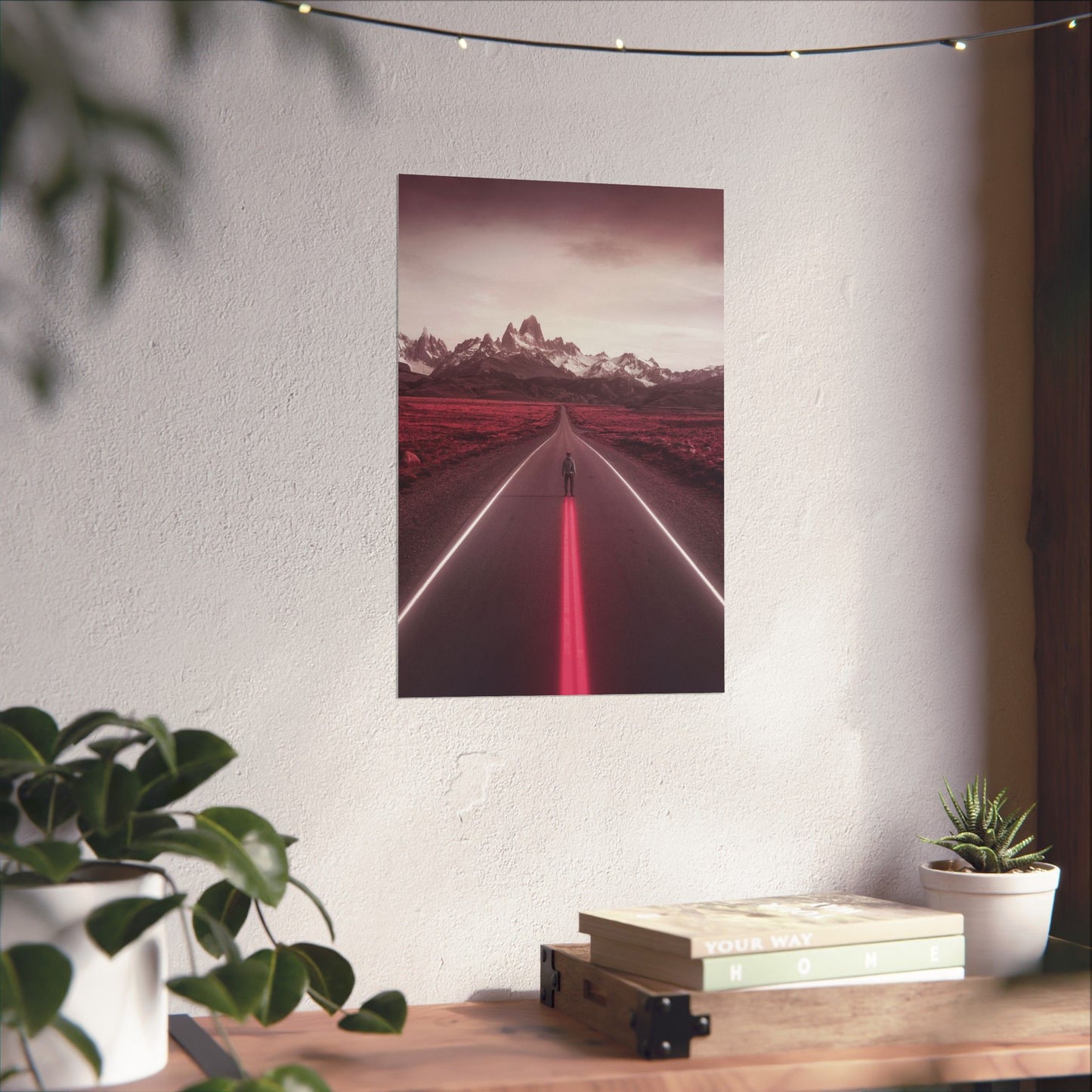 Poster of a red road with mountains on a white wall above a desk with plants and books.