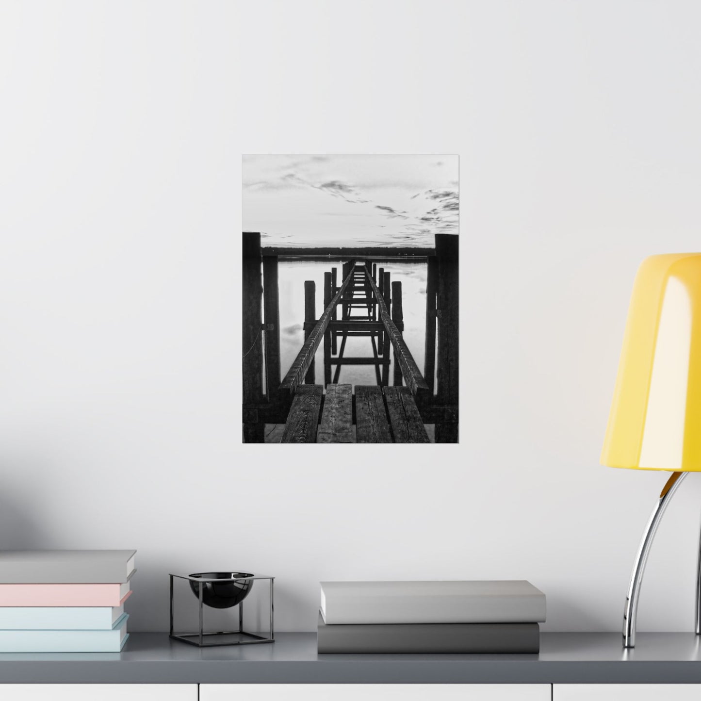 Black and white photo of a dock on a wall above a shelf with books and a lamp.