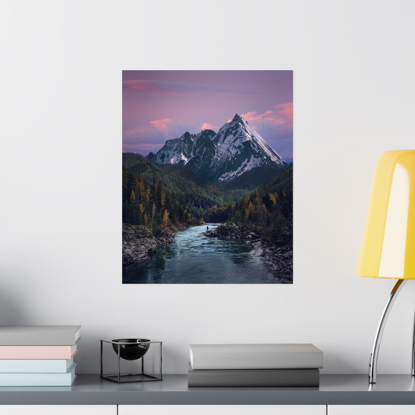 Framed mountain landscape print on a wall above a shelf with books and decor items.