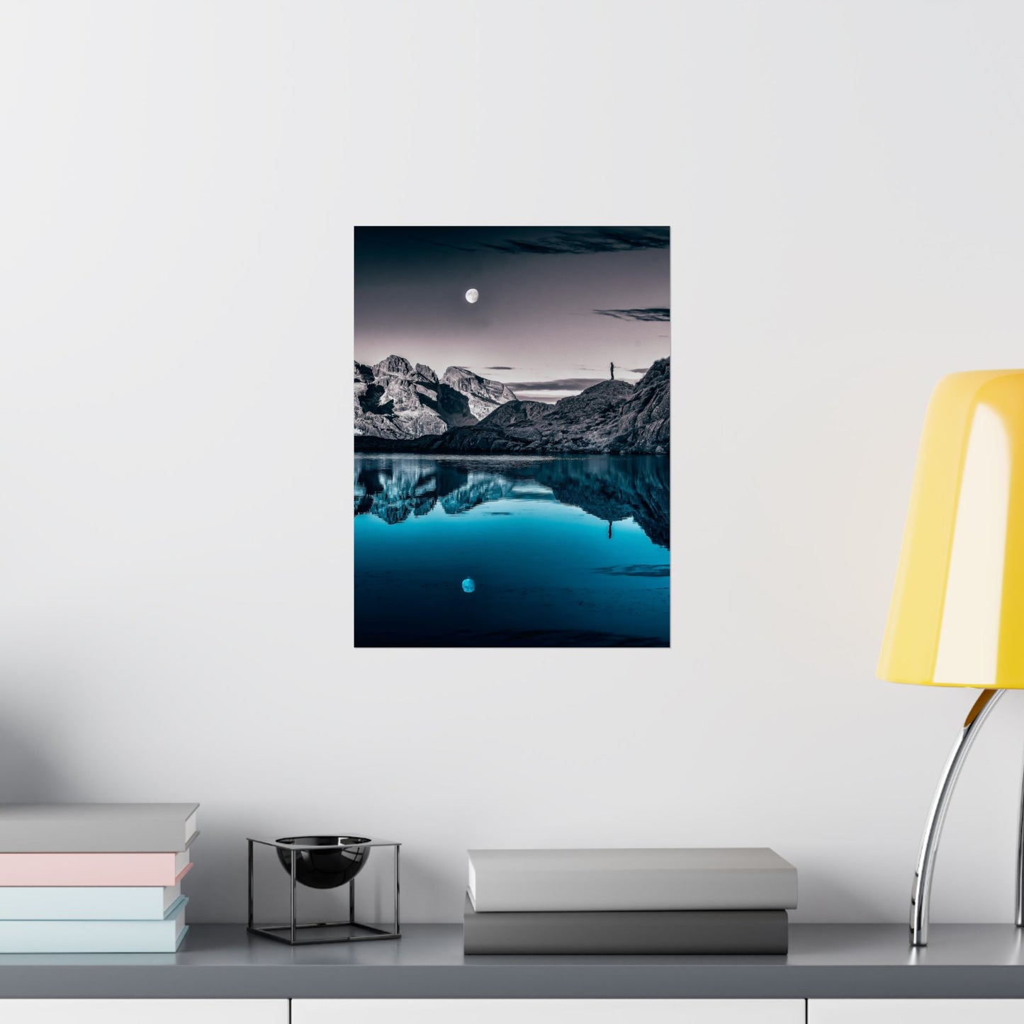 Framed mountain landscape print on a wall above a shelf with books and a lamp.