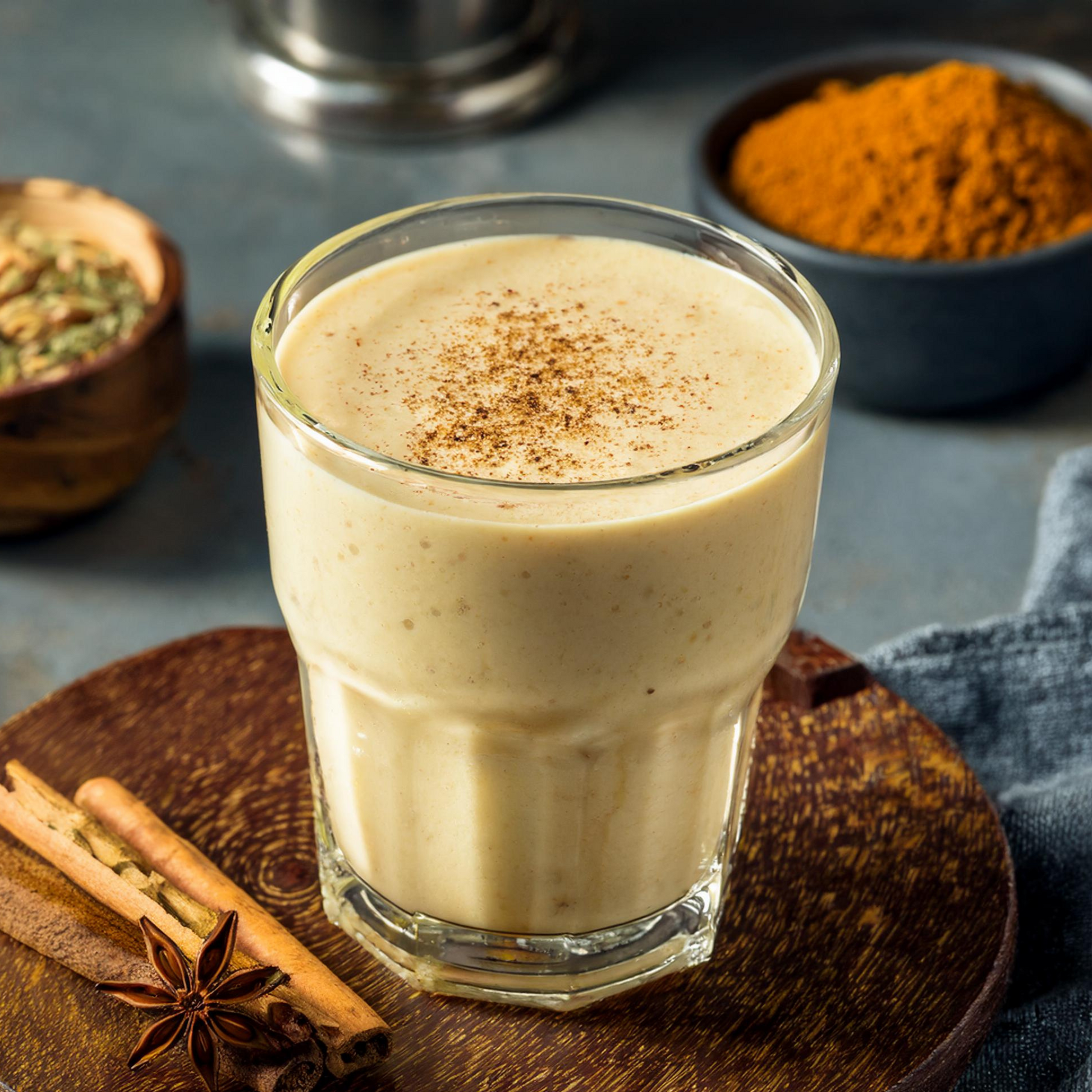 A glass of smoothie with a straw, garnished with cinnamon sticks and a sprinkle of cayenne pepper, served on a wooden table with spices and a wooden bowl in the background.