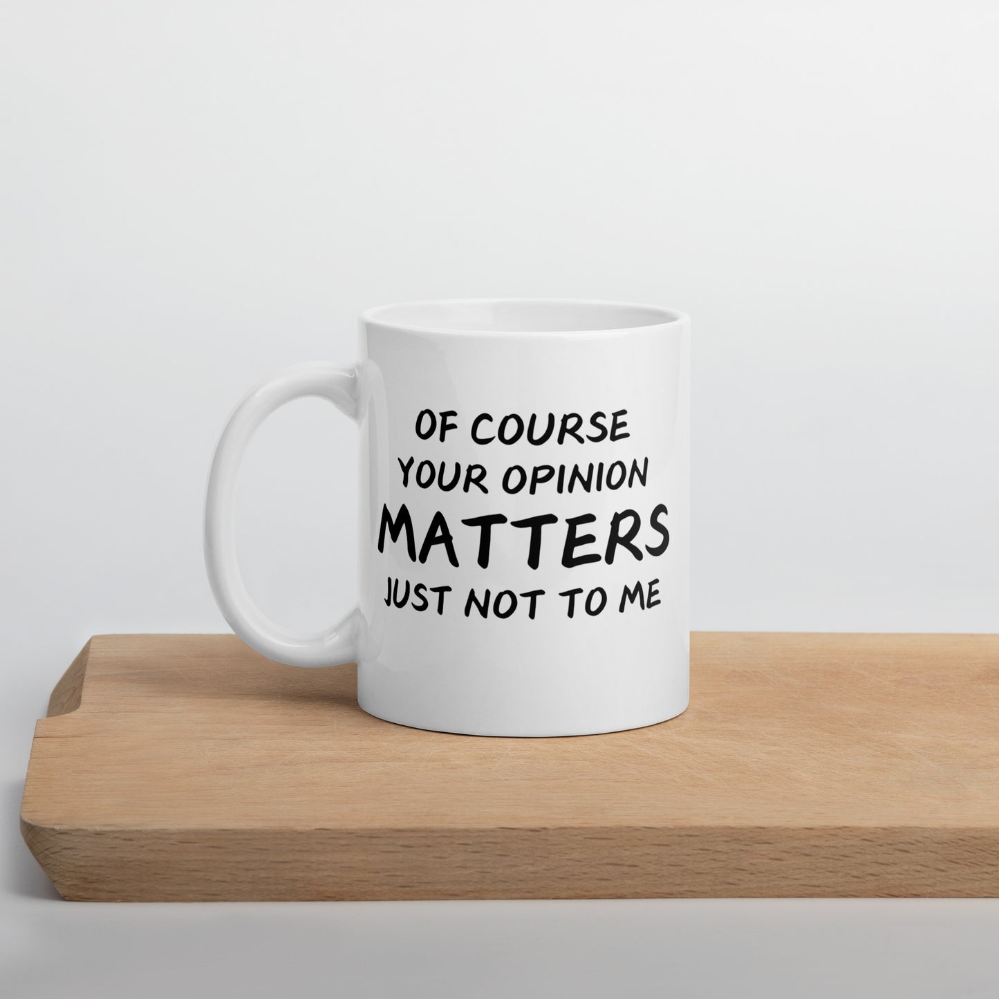 A white ceramic mug with text printed on it, placed on a wooden surface. The text reads, 'Of Course Your Opinion Matters, Just Not to Me.