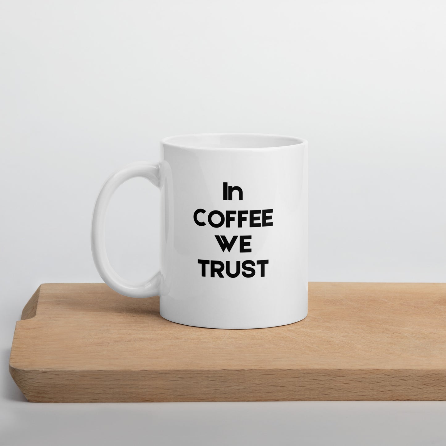 A white ceramic mug with the text 'In Coffee We Trust' printed in black on the front, placed on a wooden board.