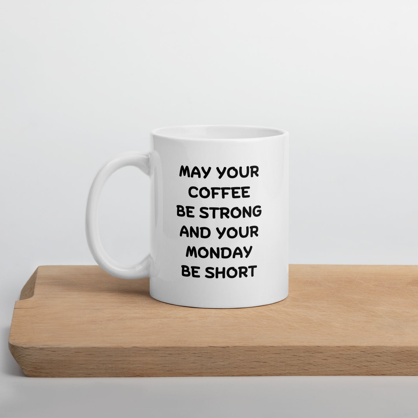 A white ceramic coffee mug with black text reading 'MAY YOUR COFFEE BE STRONG AND YOUR MONDAY BE SHORT' on a wooden surface.