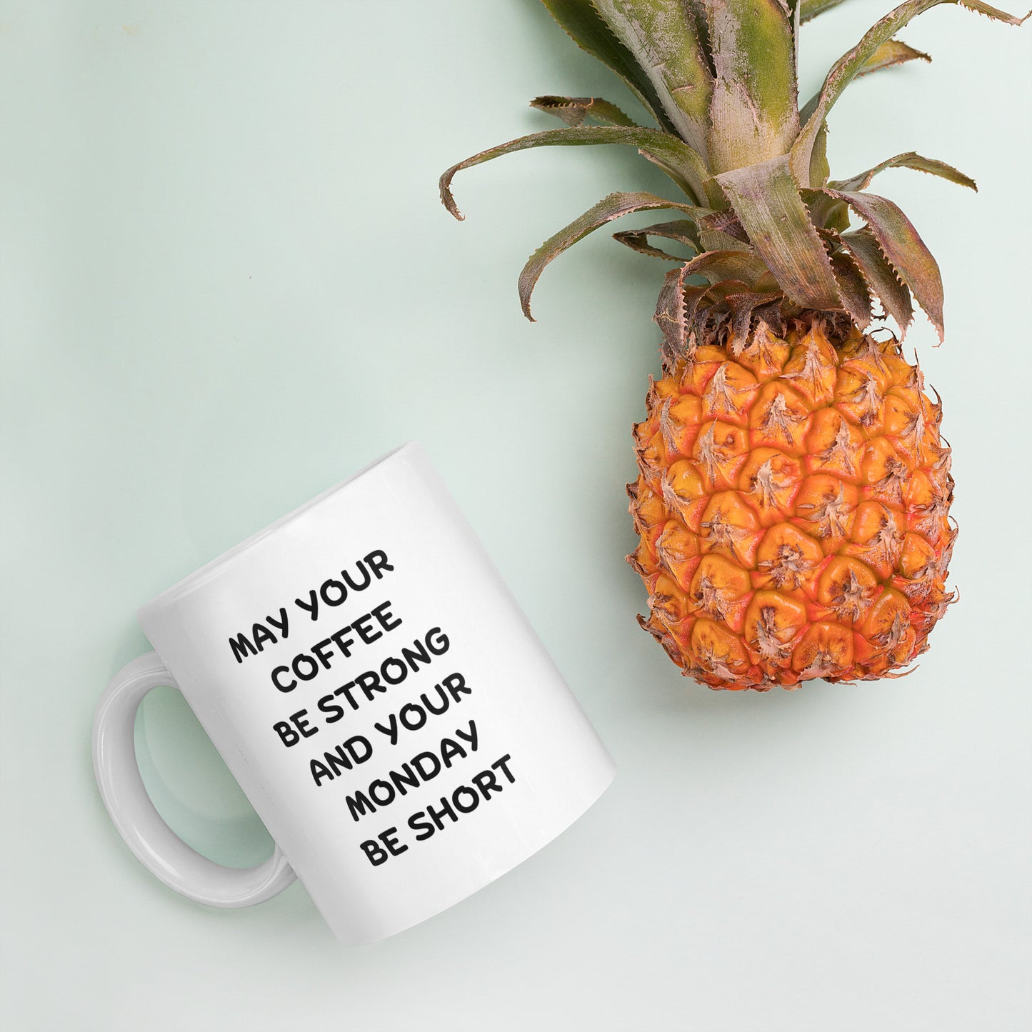A white ceramic coffee mug with black text reading 'MAY YOUR COFFEE BE STRONG AND YOUR MONDAY BE SHORT'