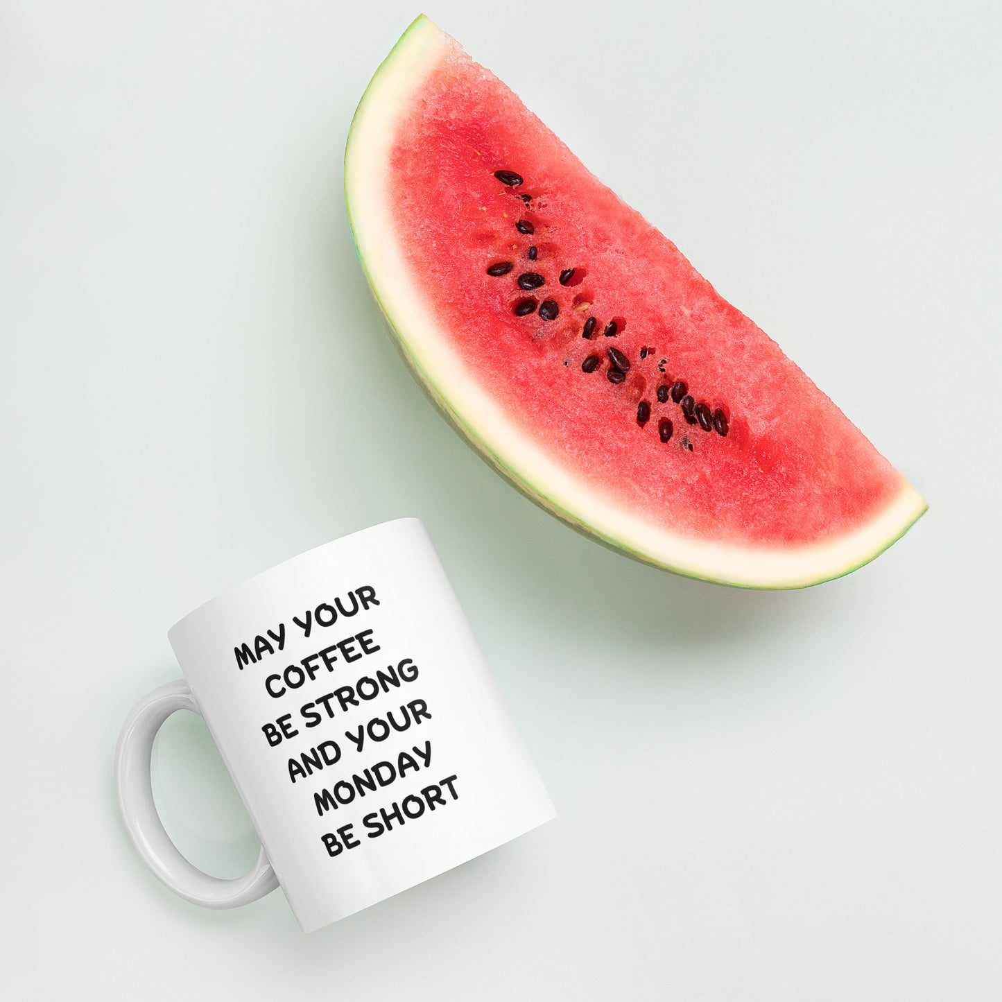 A white ceramic coffee mug with black text reading 'MAY YOUR COFFEE BE STRONG AND YOUR MONDAY BE SHORT'