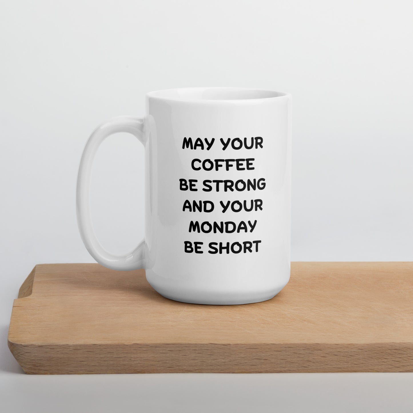 A white ceramic coffee mug with black text reading 'MAY YOUR COFFEE BE STRONG AND YOUR MONDAY BE SHORT' on a wooden surface.
