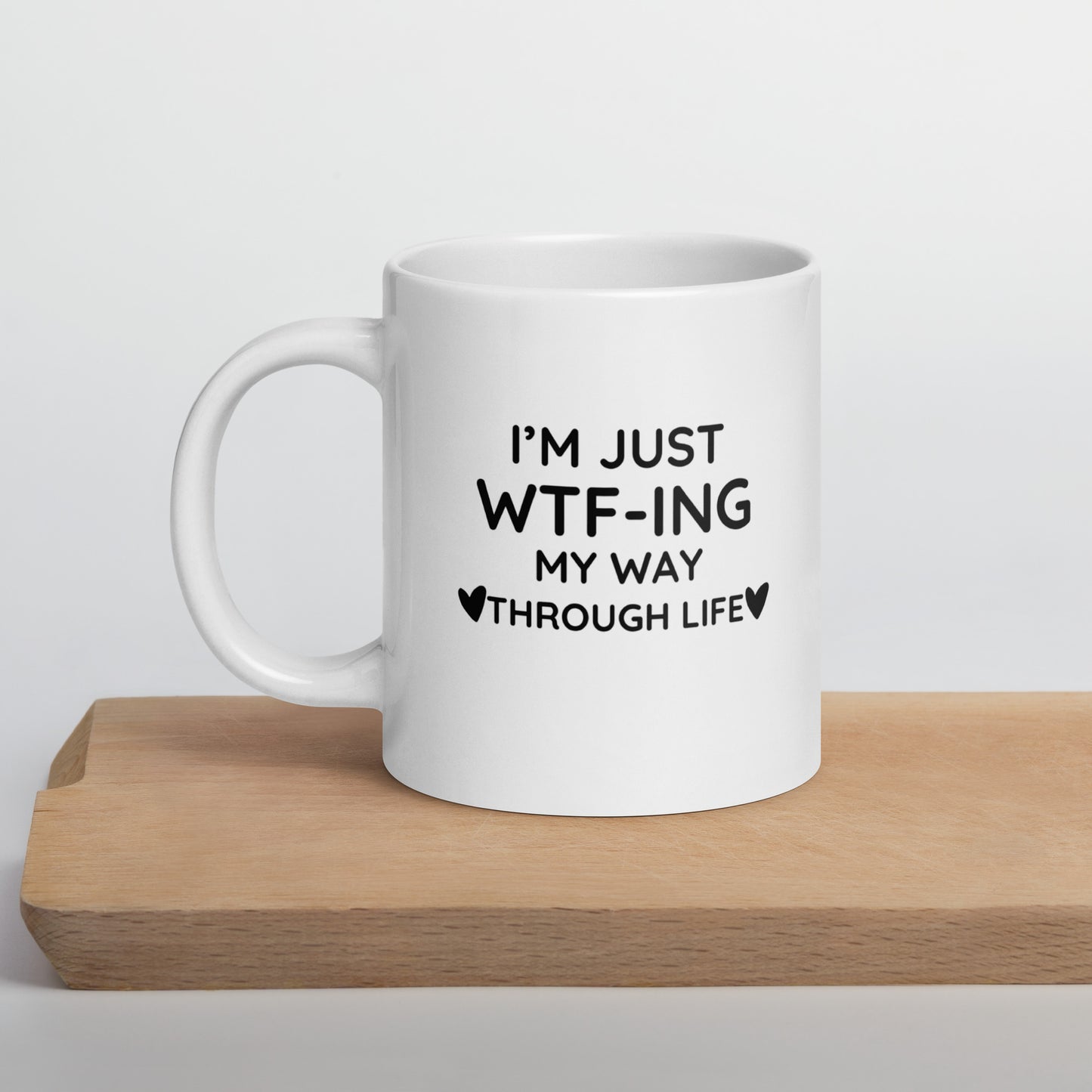 A white ceramic mug with text 'I'm just WTF-ING my way through life' printed in black color, placed on a wooden stand.