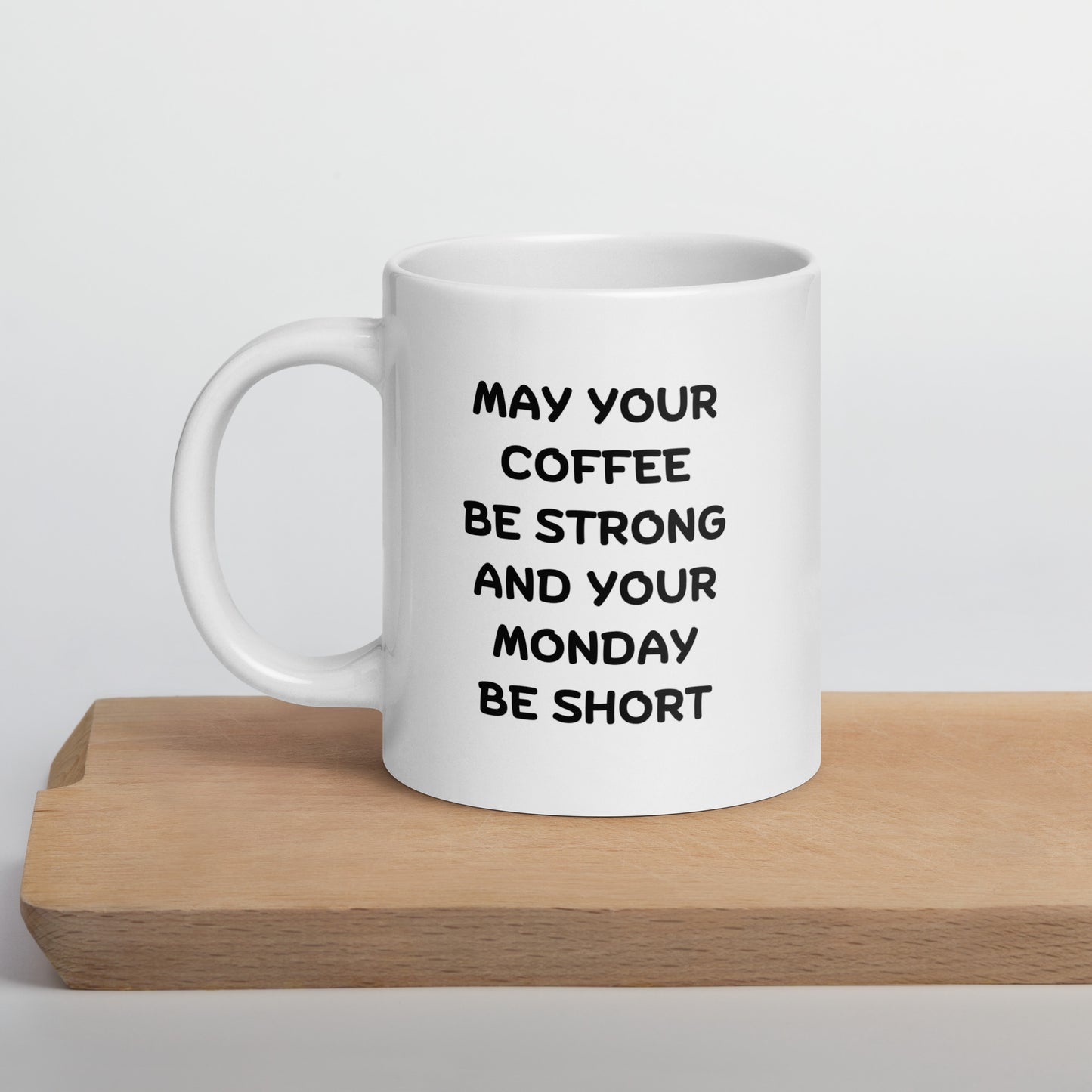 A white ceramic coffee mug with black text reading 'MAY YOUR COFFEE BE STRONG AND YOUR MONDAY BE SHORT' on a wooden surface.