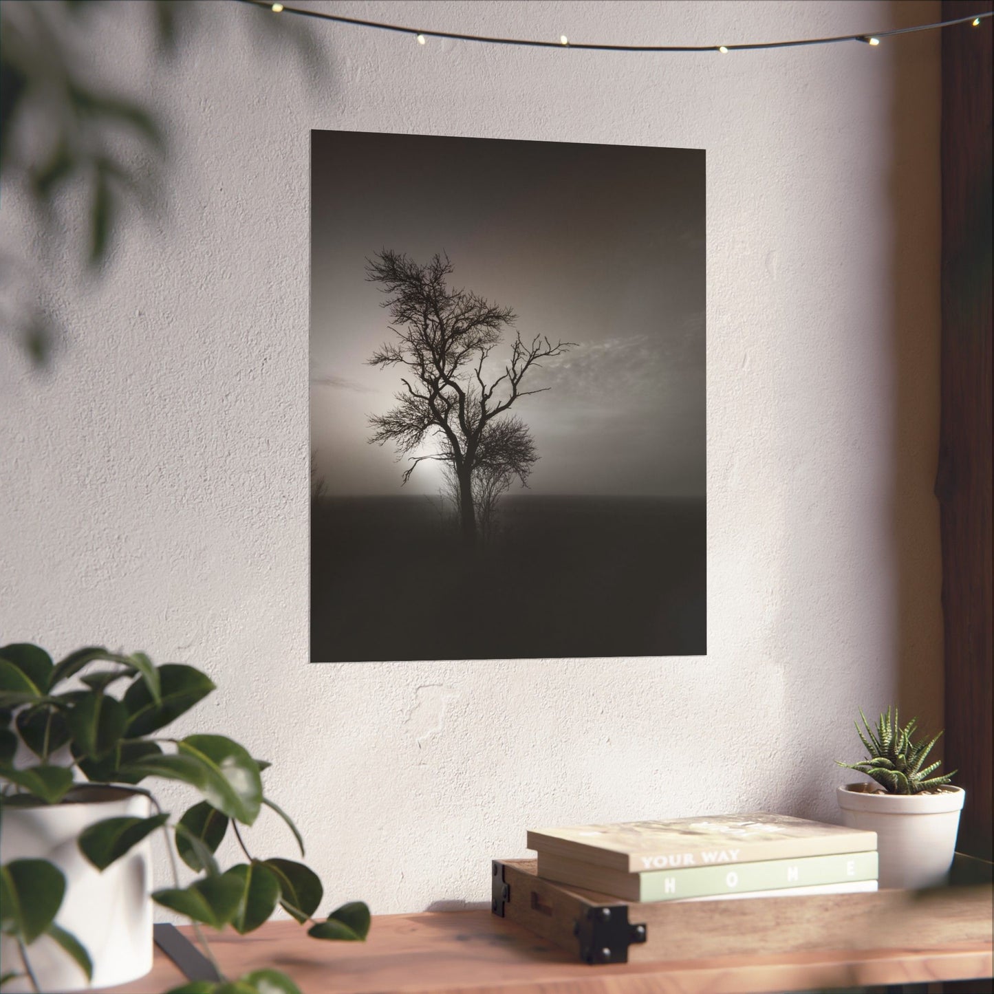 Poster of a lone tree on a foggy field hanging on a wall above a wooden desk with plants and books.