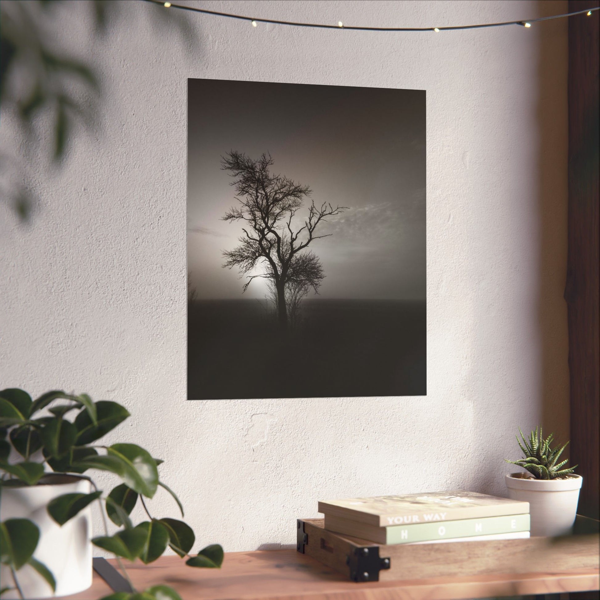 Poster of a lone tree on a foggy field hanging on a wall above a wooden desk with plants and books.