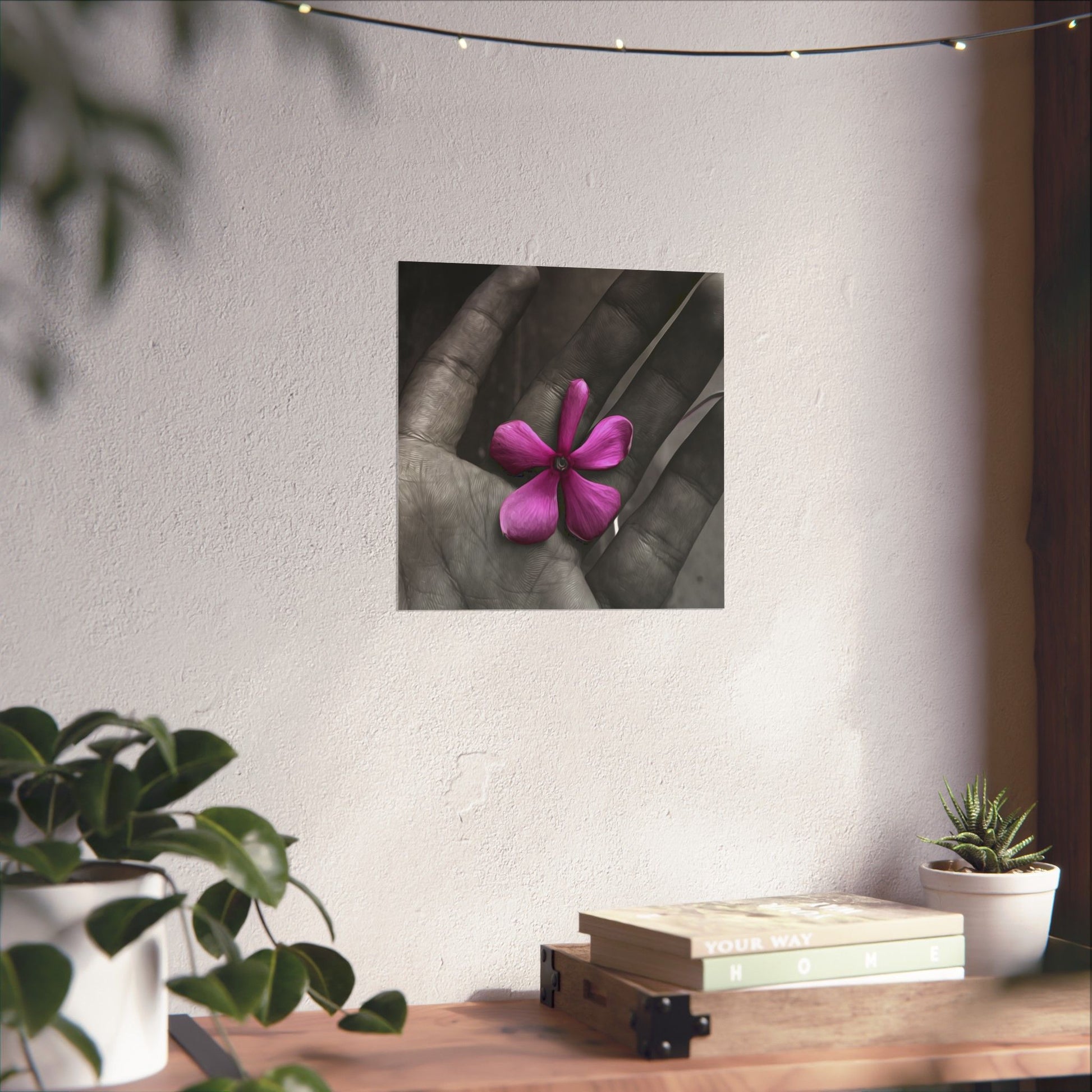 Framed artwork of a pink flower held in a hand on a wall above a wooden table with plants.