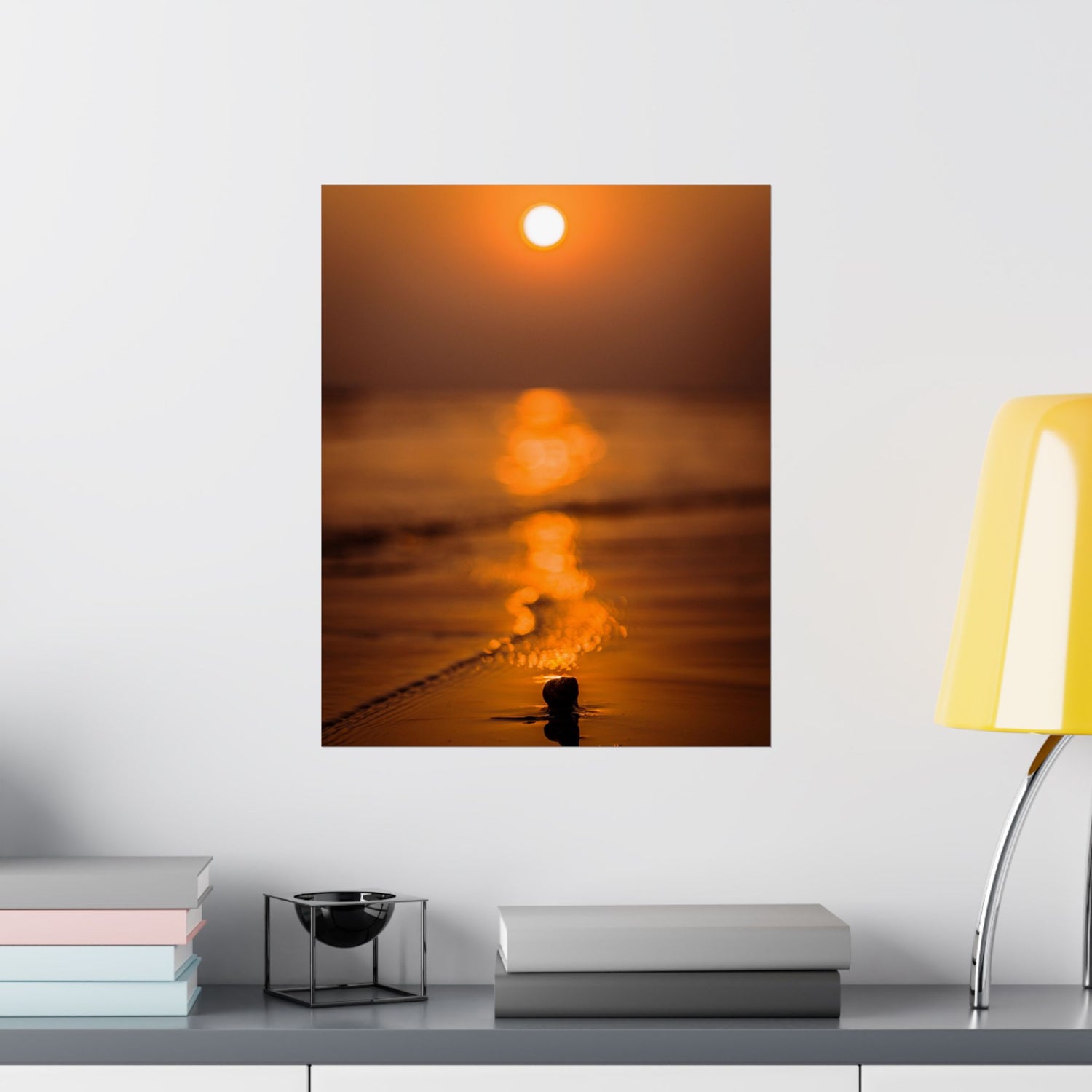 Framed artwork of a sunset over water on a wall above a shelf with books and decor.