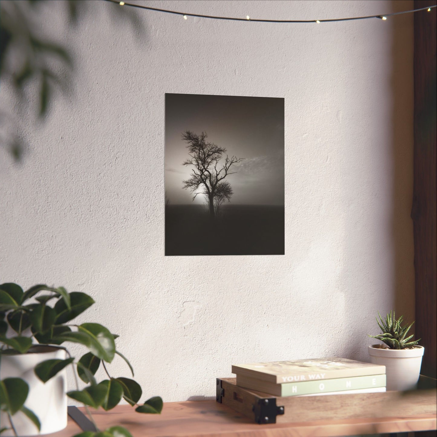 Framed black and white print of a lone tree on a wall above a wooden desk with plants.