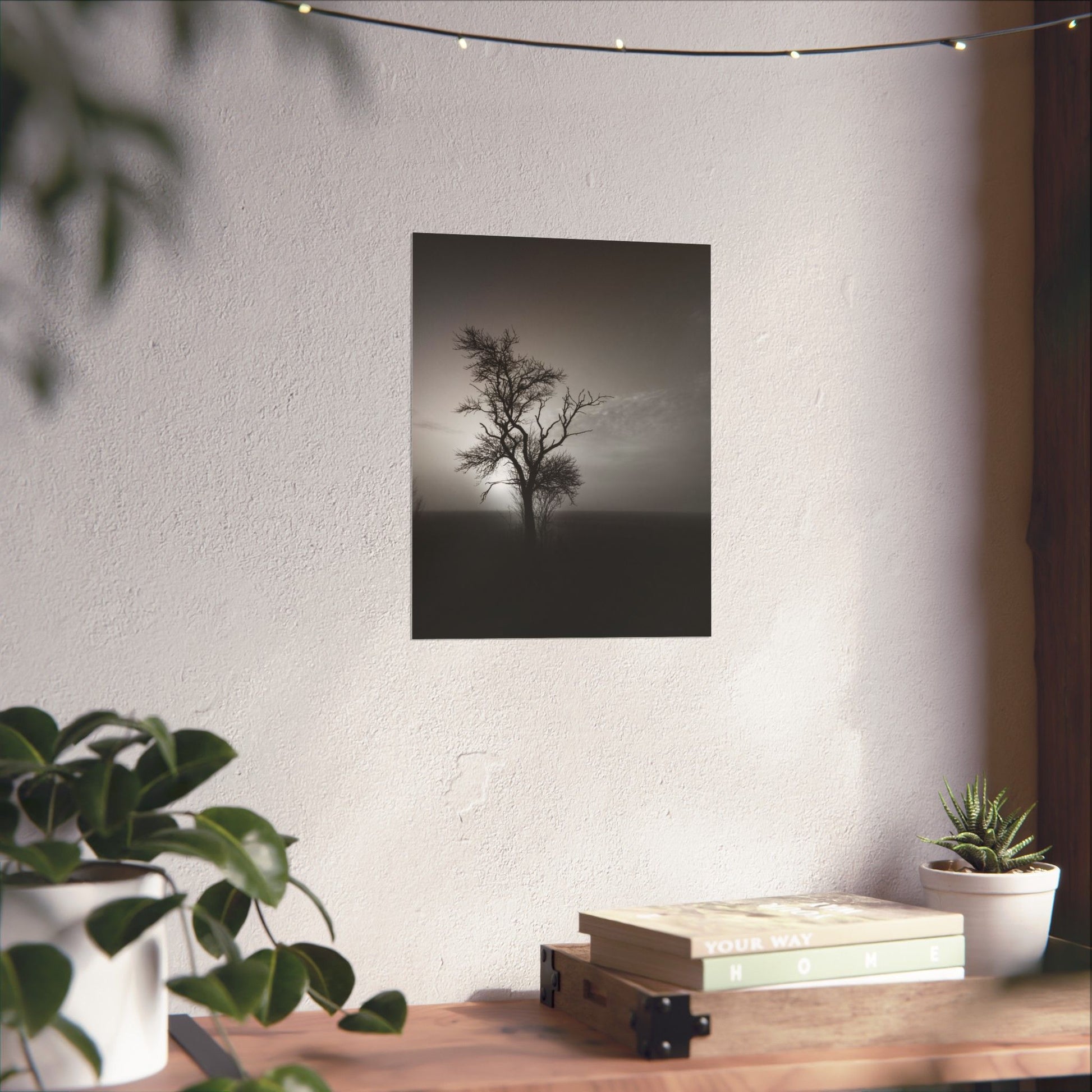 Framed black and white print of a lone tree on a wall above a wooden desk with plants.