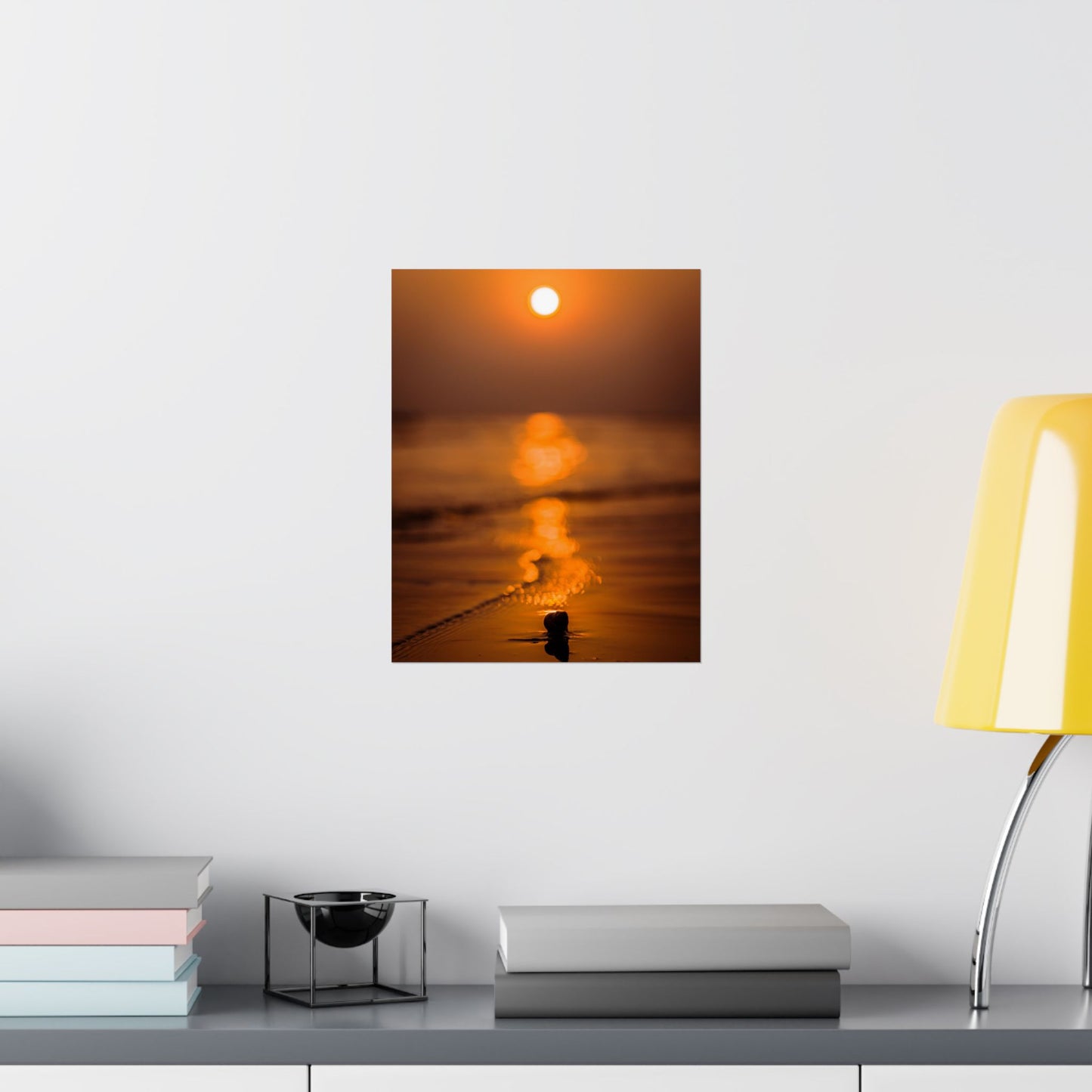 Framed artwork of a sunset over water on a wall above a desk with books and a lamp.