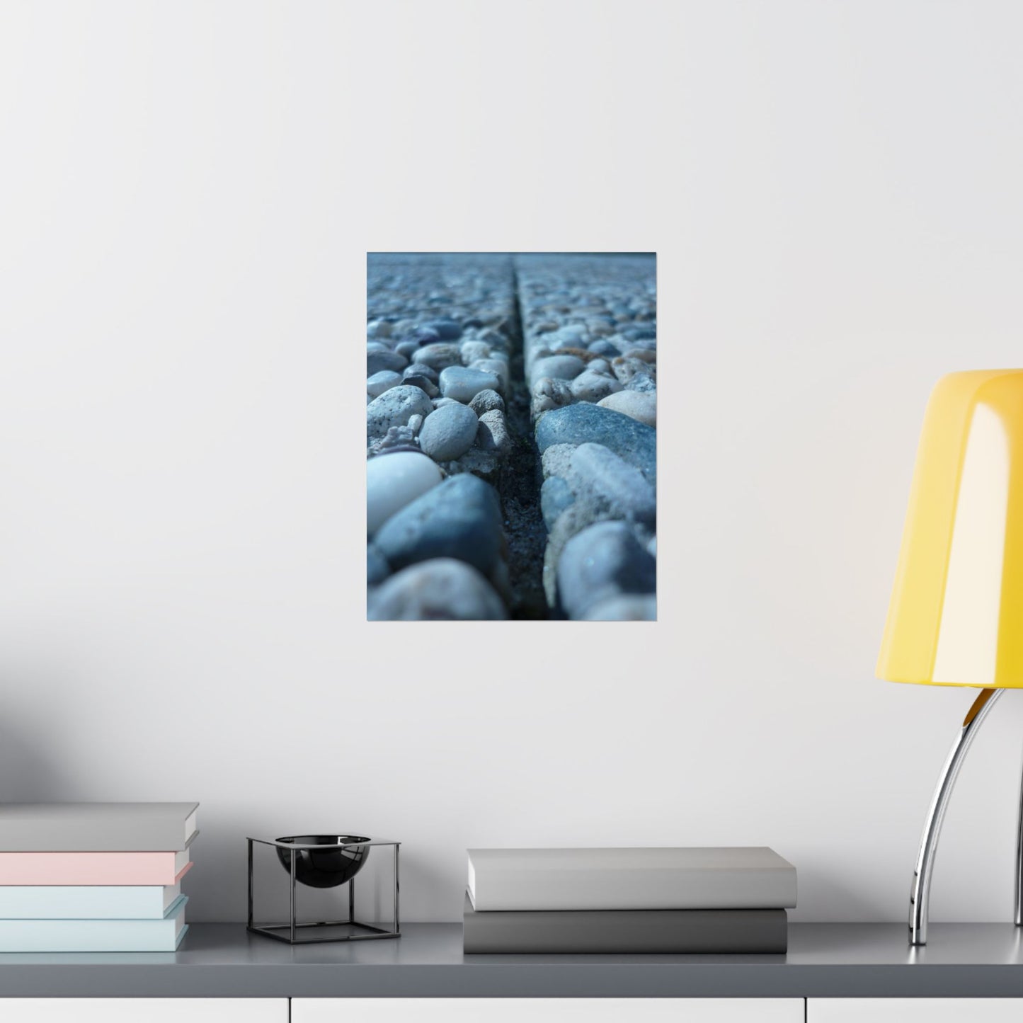 Framed artwork of textured blue surface on a white wall above a shelf with books and a lamp.