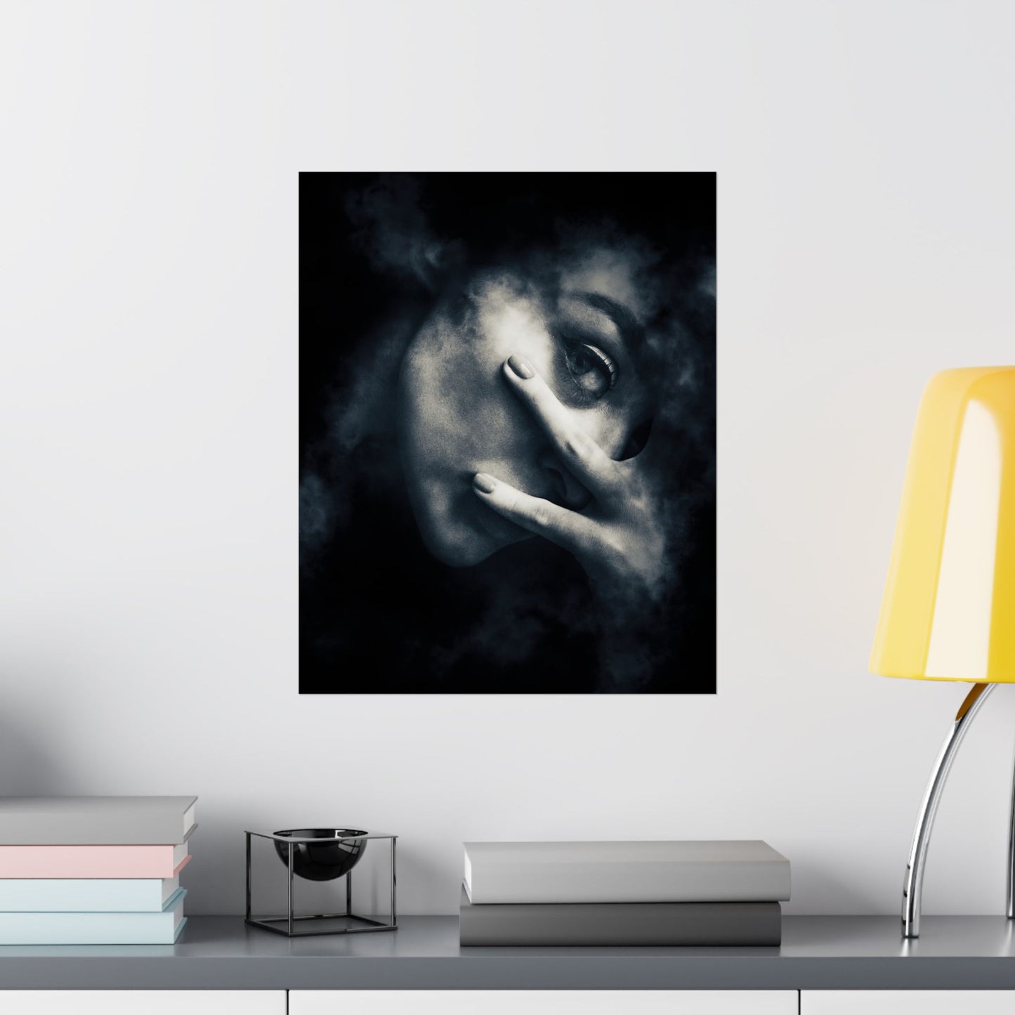 Framed artwork of a face with smoke effect on a wall above a shelf with books and a lamp.