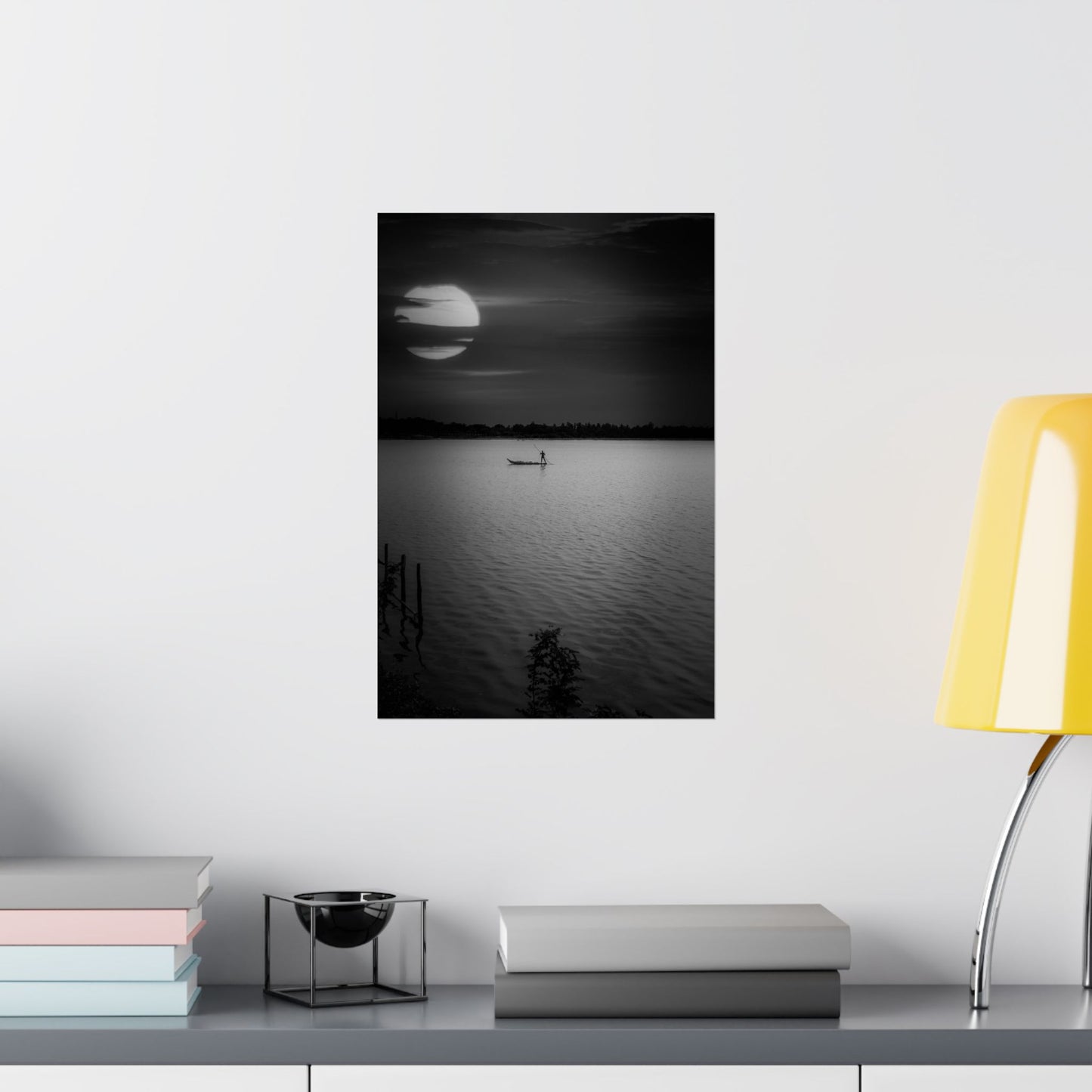 Framed black and white landscape of a sunset over water on a wall above a desk with books and a lamp.