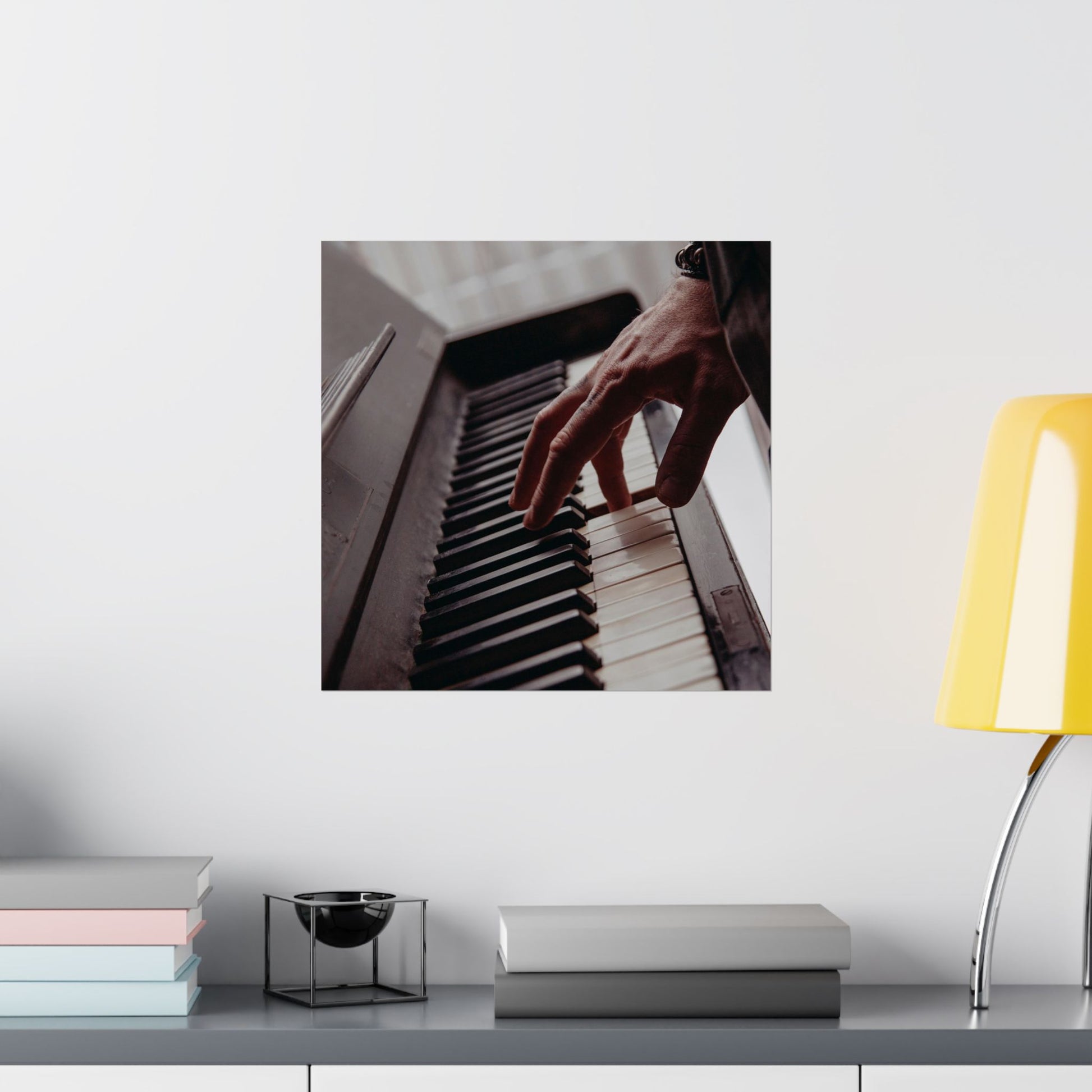 Framed picture of a hand playing piano on a wall above a desk with books and a lamp.