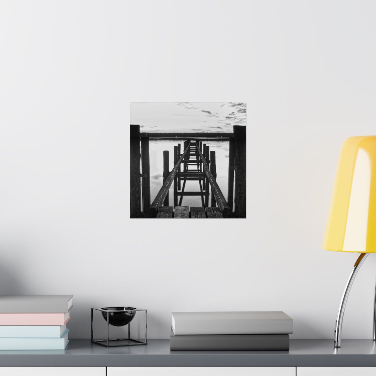 Framed black and white artwork on a wall above a shelf with books and a lamp.