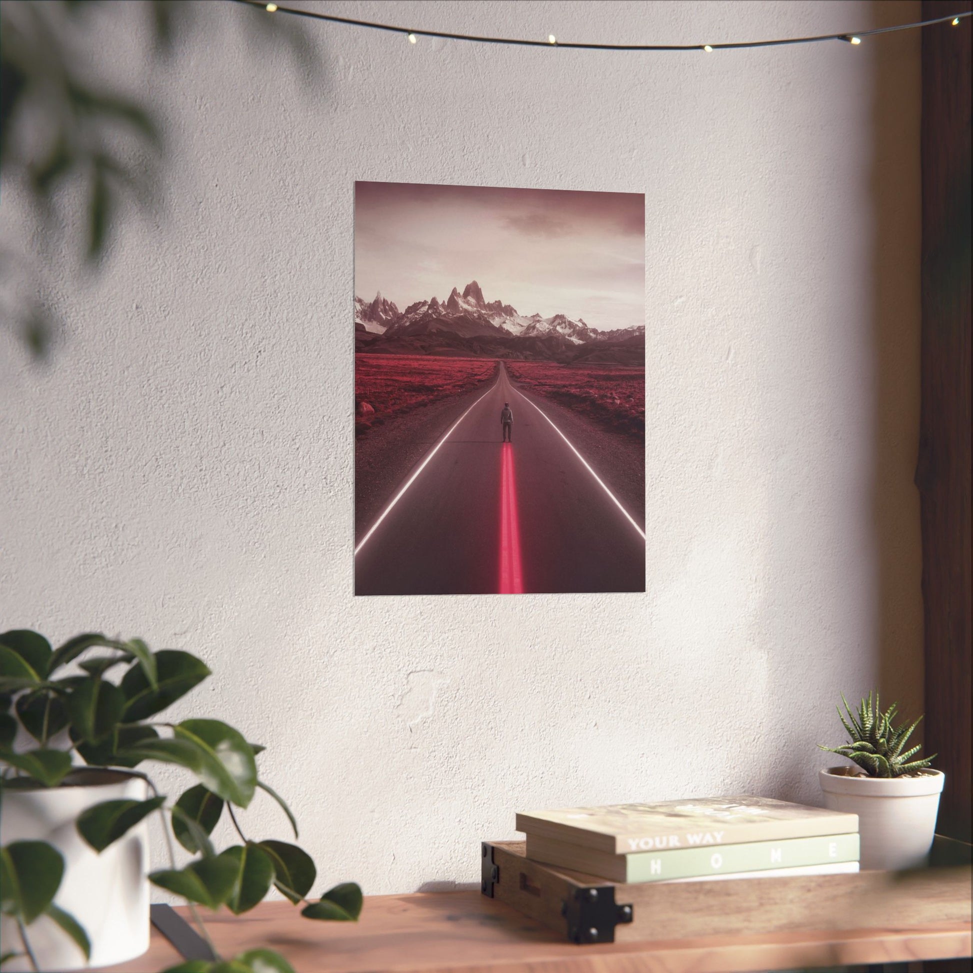 Poster of a red road with mountains on a white wall above a wooden desk with plants and books.