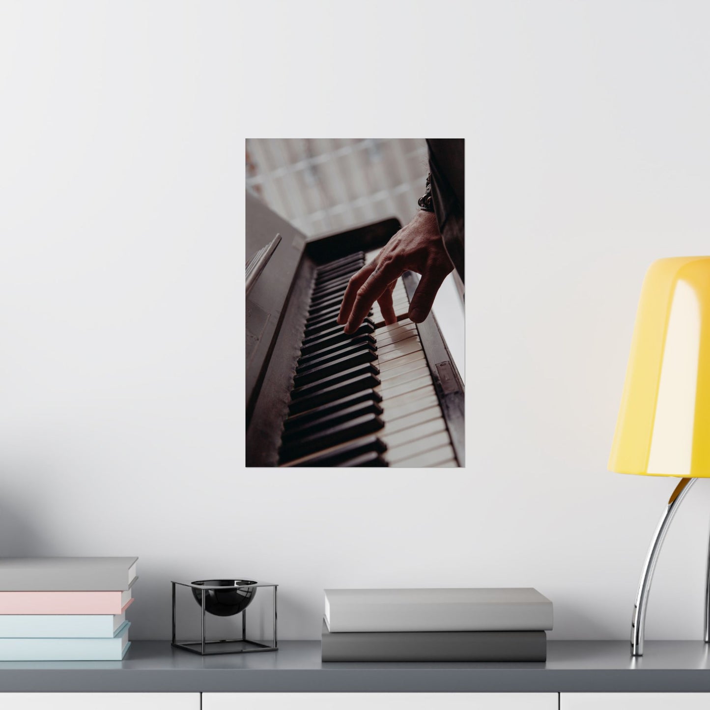 Framed picture of hands playing piano on a shelf with books and a lamp.
