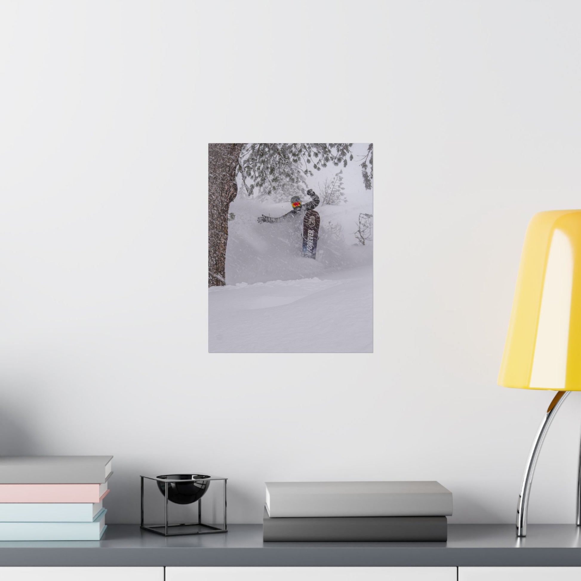 Poster of a skier in a snowy forest on a white wall above a desk with books and a lamp.