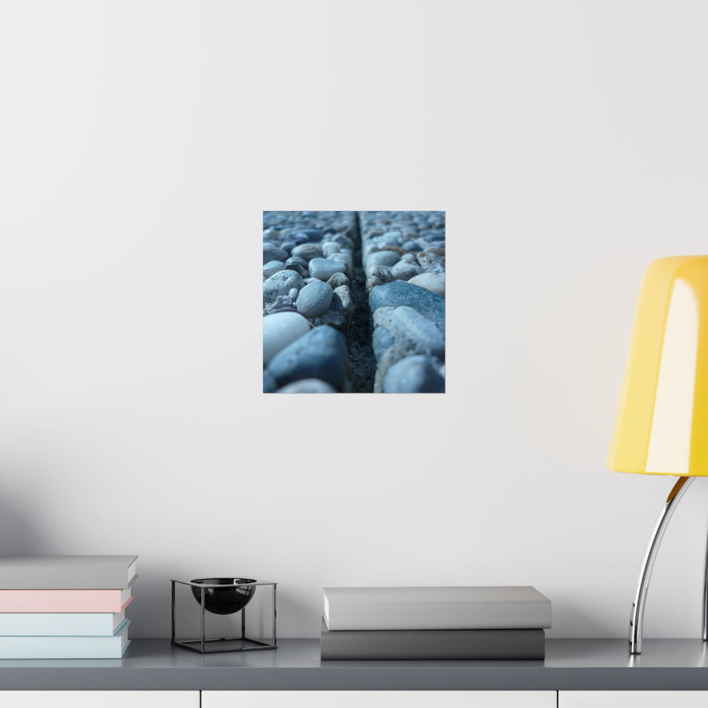 Framed artwork of pebbles on a wall above a shelf with books and a lamp.