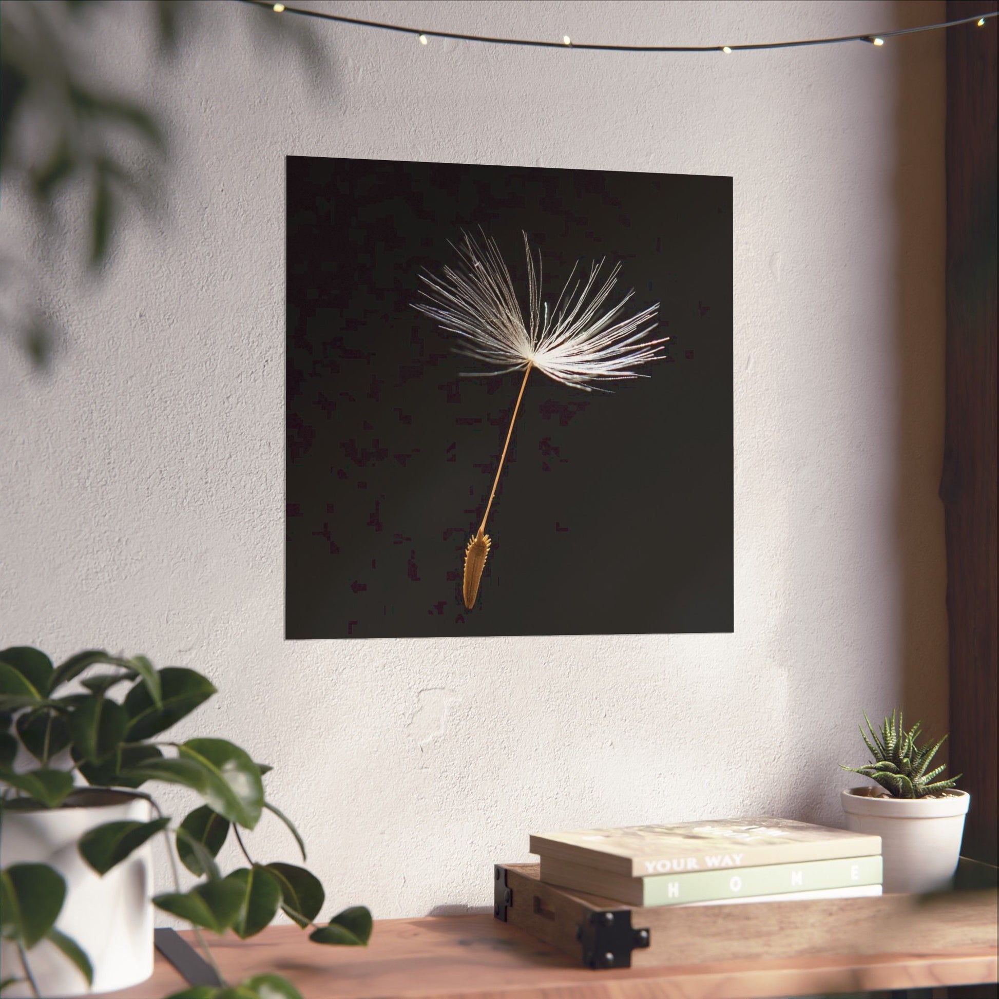 Framed artwork of a dandelion seed on a wall above a wooden table with plants and books.